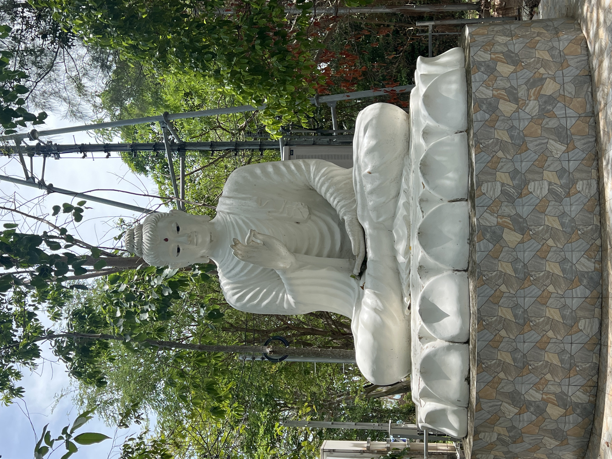 This is a medium shot, eye-level, outdoor photograph of a large, white Buddha statue situated on a tiered, mosaic-tiled pedestal in Baan Thong Krut, Thailand. The statue depicts the Buddha in a seated meditation posture, with his legs crossed and his hands resting in his lap. His eyes are closed, and a small red dot is visible on his forehead. The statue is made of smooth, white material, possibly ceramic or painted concrete, and is adorned with flowing robes.
The pedestal is circular and rises in two sections. The lower section is covered in a mosaic of irregularly shaped tiles in shades of grey, beige, and terracotta, creating a geometric pattern. The upper section of the pedestal is a lotus-flower-shaped base, upon which the Buddha statue sits.
Behind the statue, lush green trees and foliage create a natural backdrop. A tall, metal utility pole with various equipment attached is visible to the right of the statue, interspersed with the greenery. The sky above is a pale blue with scattered white clouds. The lighting suggests it is daytime, with sunlight filtering through the leaves, casting shadows on the statue and its base. There are no people visible in the image. The scene appears to be in a tranquil, possibly temple or shrine environment.