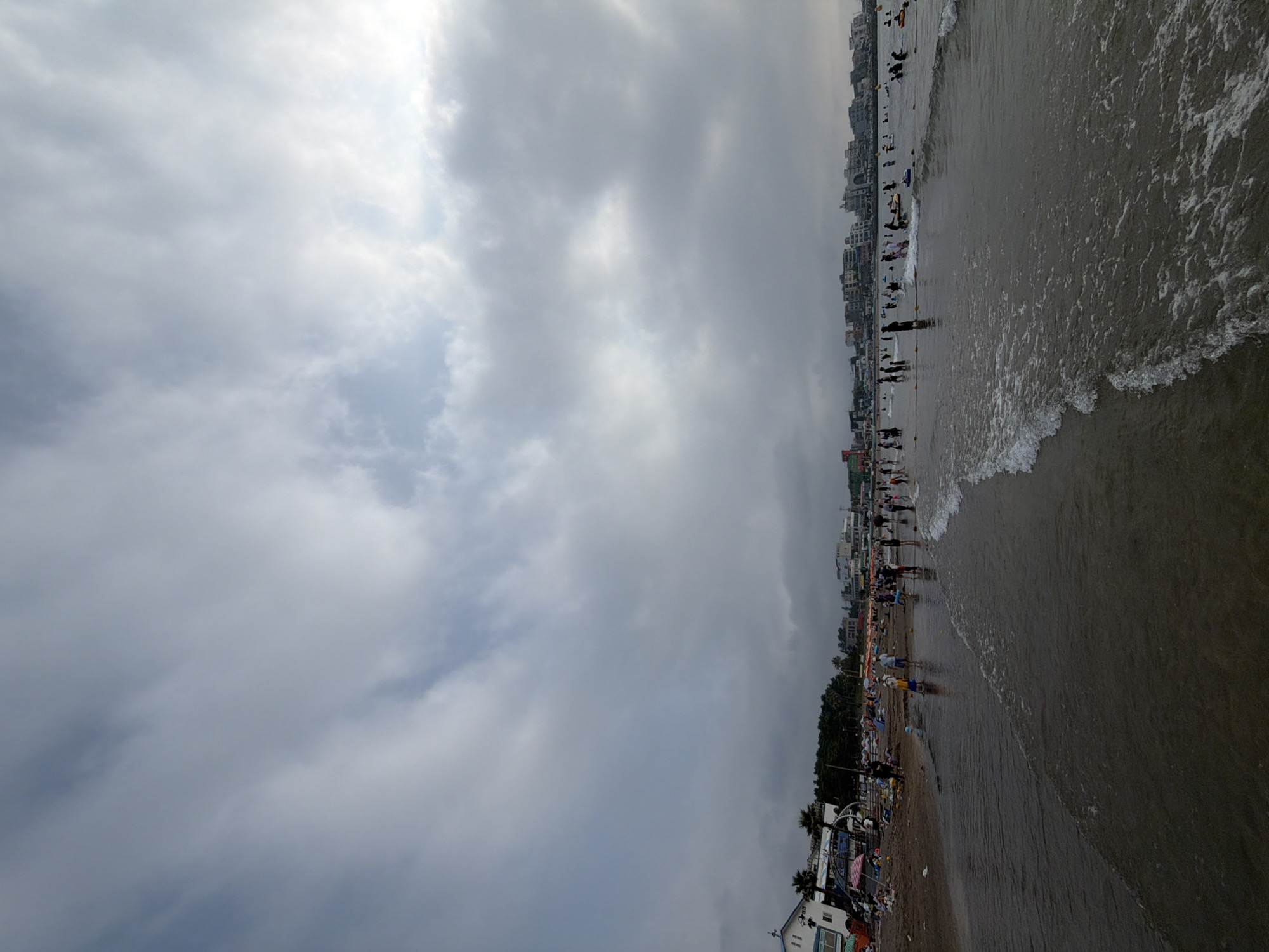 This is a wide-angle, low-angle, outdoor, full-shot photograph of a crowded beach scene on Jeju Island, South Korea, under a cloudy sky. The image is dominated by a vast expanse of overcast sky, with dramatic, textured clouds covering most of the upper frame. The lower portion of the image features a dark, wet sandy beach with numerous people engaged in various activities.
In the foreground, gentle waves with foamy crests are washing ashore, creating a glistening, reflective surface on the wet sand. Several individuals are visible wading in the shallow water or standing near the water's edge.
Further back on the beach, a multitude of people are scattered across the sand. Some are standing, while others appear to be sitting or interacting. Beach umbrellas and tents are visible, suggesting recreational use of the area. A few palm trees stand amongst the buildings that line the background.
The mid-ground and background reveal a strip of buildings, likely shops, restaurants, and accommodations, typical of a coastal resort town. These structures are of varying heights and architectural styles, creating a dense urban edge to the beach.
In the water, beyond the immediate shoreline, more people can be seen, some swimming, others potentially engaged in water sports like paddleboarding, as indicated by the presence of boards in the water. The overall atmosphere appears to be one of a lively, popular beach destination despite the overcast weather.