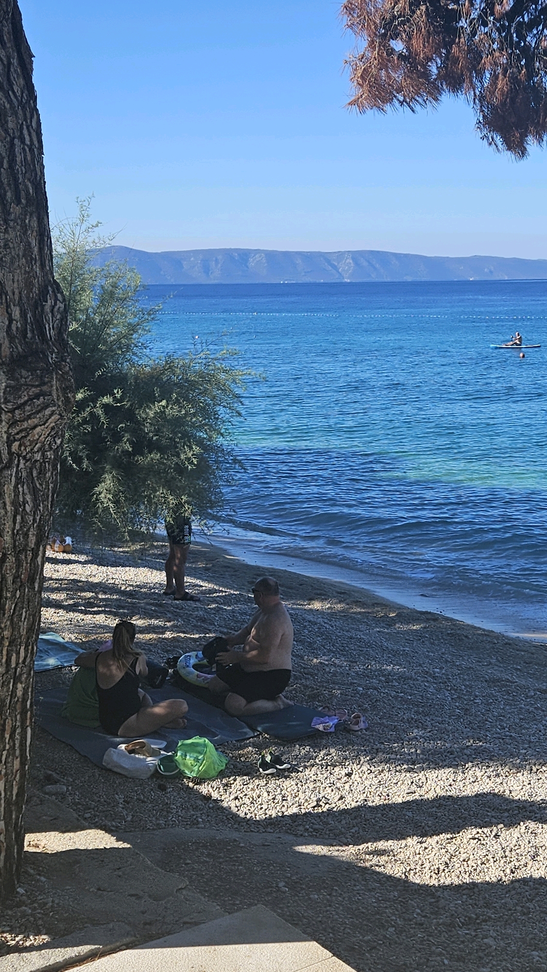 In Općina Tučepi, Croatia, on a bright, sunny day, a pebble beach stretches along the tranquil turquoise sea. The water is calm, with distant mountains forming a hazy blue backdrop. In the foreground, under the dappled shade of a large tree, two individuals are relaxing on the beach. A woman in a black swimsuit is seated on a mat, facing away from the camera, with a green towel draped over her. Beside her, a shirtless man with dark hair is kneeling on another mat, holding a black object. Scattered around them are beach essentials: a yellow and white object, a bright green bag, and a pair of black sneakers. To the right, a pink and purple child's sandal rests on the pebbles. Further back, a person stands near a lush green tree, and in the distance, a single figure is visible on a paddleboard in the water. The scene conveys a sense of peaceful leisure on a Mediterranean coastline.
