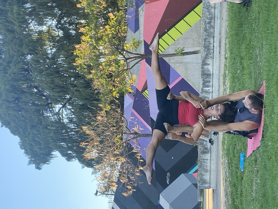 The image captures an outdoor acro-yoga session in a public park setting in the Palermo neighborhood of Buenos Aires, Argentina, likely within the Bosques de Palermo or Paseo de la Infanta area, identifiable by the characteristic urban green spaces and geometric street art.
In the foreground, a male individual, acting as the 'base', lies on his back on a pink yoga mat. He wears a dark grey t-shirt and dark shorts. His feet support the hips/upper thighs of a female 'flyer' who is inverted above him, while his hands grasp her hands. The female flyer wears a red tank top and black athletic shorts, with her legs extended widely in a horizontal straddle split. She is barefoot and smiles downward towards the base. A light blue water bottle is visible on the grass to the left of the yoga mat.
The mid-ground features a concrete retaining wall covered in vibrant geometric graffiti, utilizing angular shapes and colors including purple, pink, black, green, and yellow. Behind this wall, various trees are present; a large, dark green weeping tree dominates the upper background, while smaller trees, some deciduous, are positioned in front of the wall. A clear, light blue sky is visible at the top. On the far left and right edges, partial views of other individuals indicate a public gathering or group activity. A black streetlamp is visible in the upper left, behind the graffiti wall. The scene is illuminated by natural light, suggesting either early morning or late afternoon.