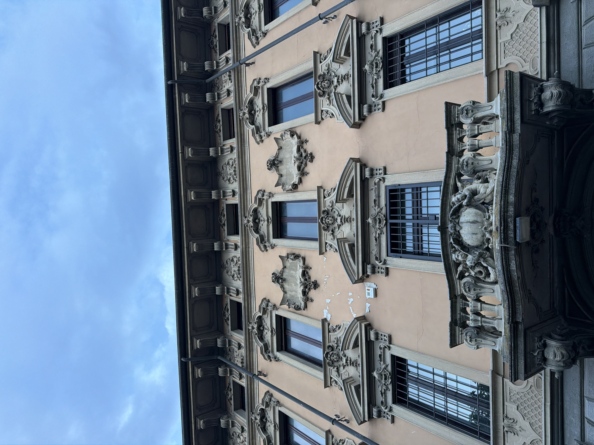 A low-angle, full-frame shot captures the ornate facade of a peach-colored building under a cloudy sky. The building features multiple windows with dark frames and grates, some of which reflect the sky. Each window is adorned with elaborate stone carvings, including faces, scrolls, and decorative patterns. A prominent balcony, also made of stone, juts out from the facade, decorated with cherubic figures and a central globe. The building's architectural style is rich with ornamentation, suggesting historical significance. A dark downspout runs vertically along the facade. The image is taken in Salò, Italy.