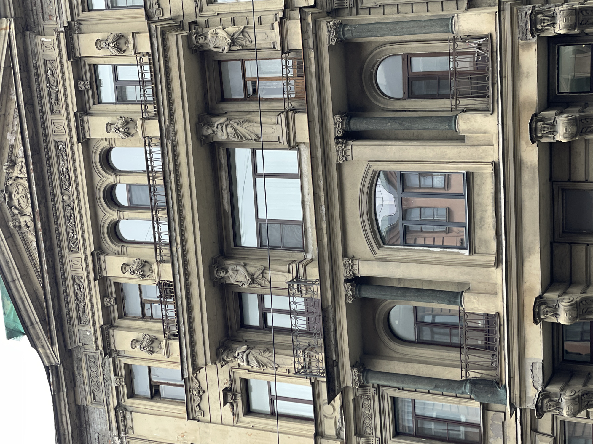 This is an upward-angled shot of the facade of the Eliseyev Emporium building in Saint Petersburg, Russia. The building has an ornate, historical architectural style featuring multiple windows, columns, and decorative elements. Some windows are rectangular while others are arched. The facade has detailed carvings, including sculptures of figures and decorative patterns. The building is made of a light-colored stone material. The sky is visible through some of the windows, suggesting an overcast day.