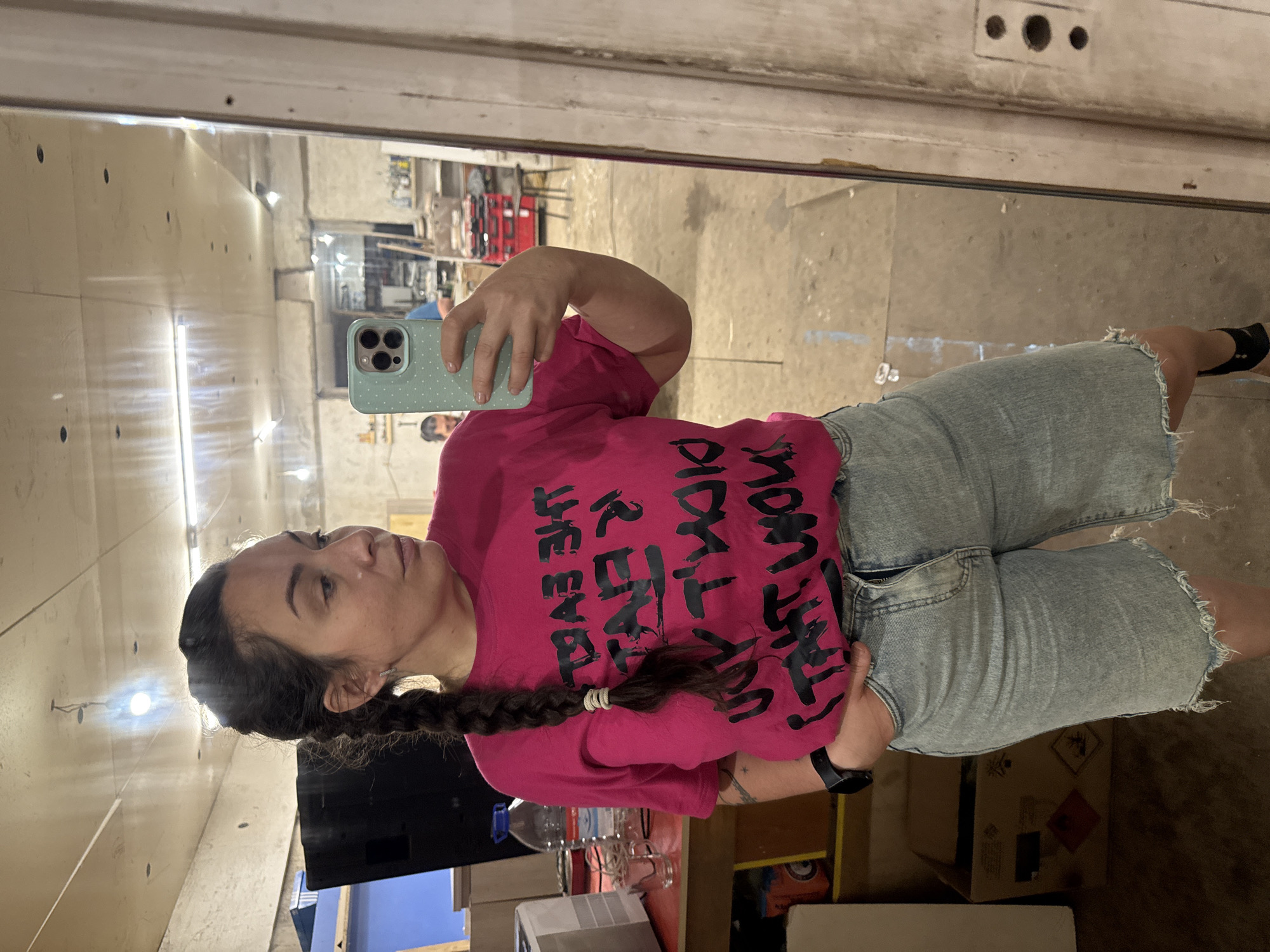 The media file is a self-portrait taken in a mirror. The main subject is a young woman with dark hair in a braid, wearing a bright pink t-shirt with black text and light blue denim shorts. She is holding a smartphone in her left hand, angled to capture her reflection. Her expression is neutral, looking at her reflection in the mirror.
The setting appears to be an indoor space, possibly a workshop or garage, given the concrete floor, exposed lighting fixtures, and various objects reflected in the mirror. These reflections show shelves with items, boxes, and what looks like some machinery or equipment in the background. The lighting is artificial, casting bright reflections on the surfaces.
The text on the woman's t-shirt is in a stylized, bold font. Some of the visible words appear to be "FIGHT", "THE", "VOICE", and possibly "PAUSE". The exact phrase or meaning is not entirely clear due to the angle and style of the lettering.
There are no discernible cues for the time of day or weather. The overall impression is a casual, candid moment captured by the subject herself in what seems to be a utilitarian environment.