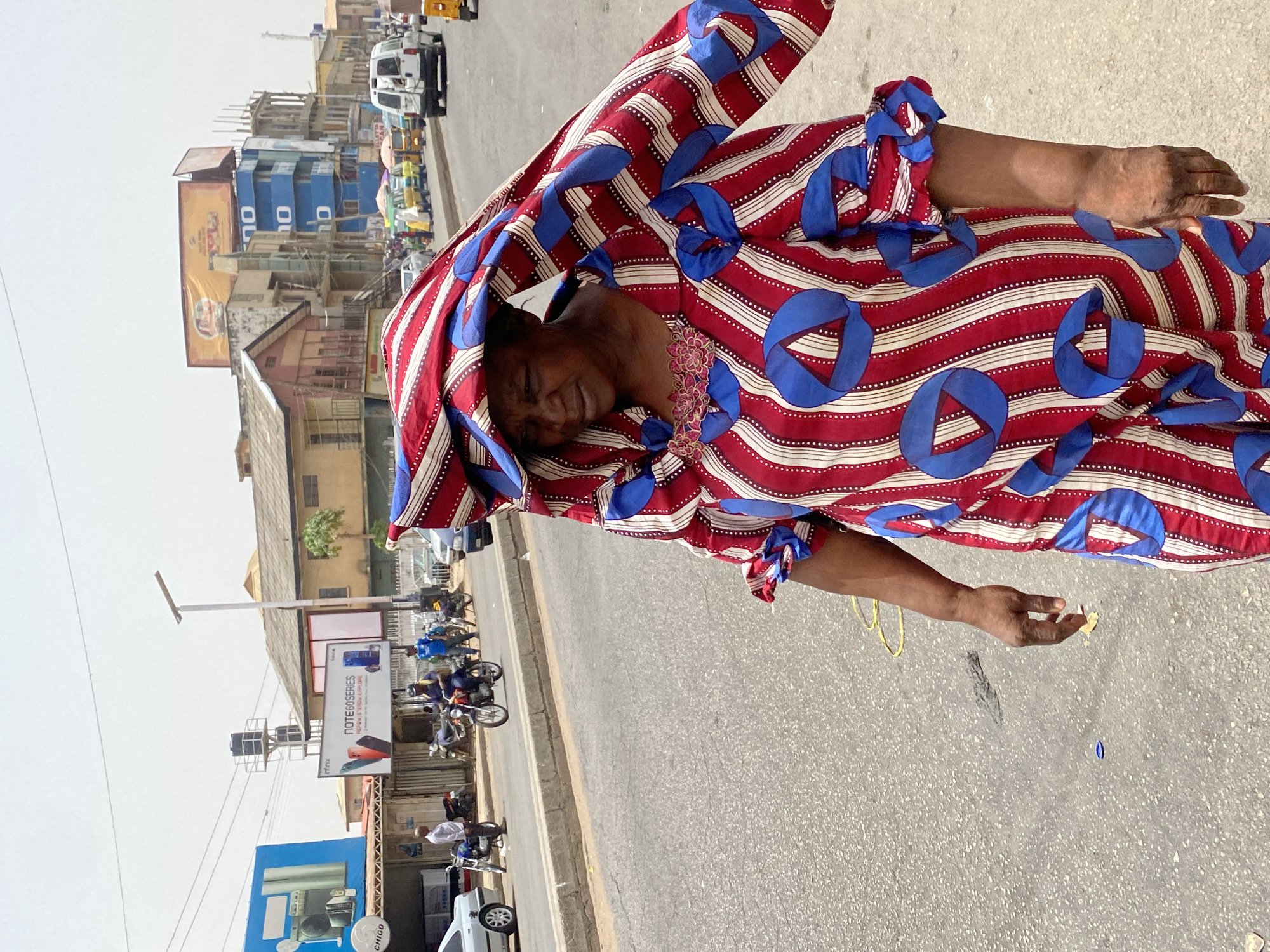 A woman stands on the street of Maiburiji, Nigeria. She wears a red, white, and blue patterned garment. The street is paved with asphalt. Buildings, billboards, motorcycles, and other vehicles populate the background of the scene.