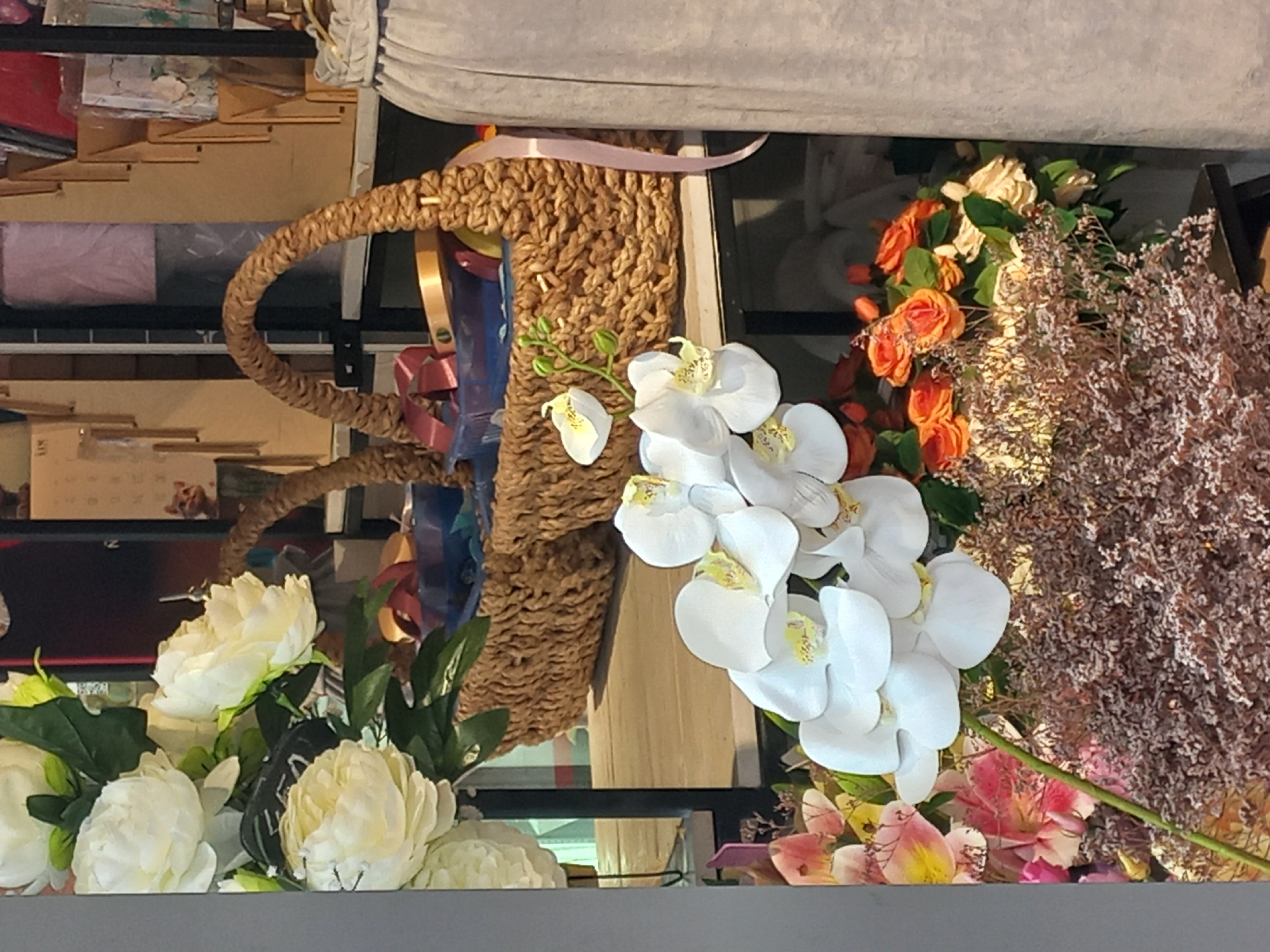 The image depicts a close-up view of a floral arrangement, seemingly indoors. A woven basket with a pink ribbon sits beside a display of flowers, including white orchids and what appear to be roses in various colors like orange and white. Additionally, a dried floral element with a textured, brown appearance is present. The arrangement is on a shelf or display, with glimpses of a shelf structure behind it. The overall setting does not provide explicit geographic details, and thus it's not possible to identify Sochi, Russia, as the location based on visual elements alone.