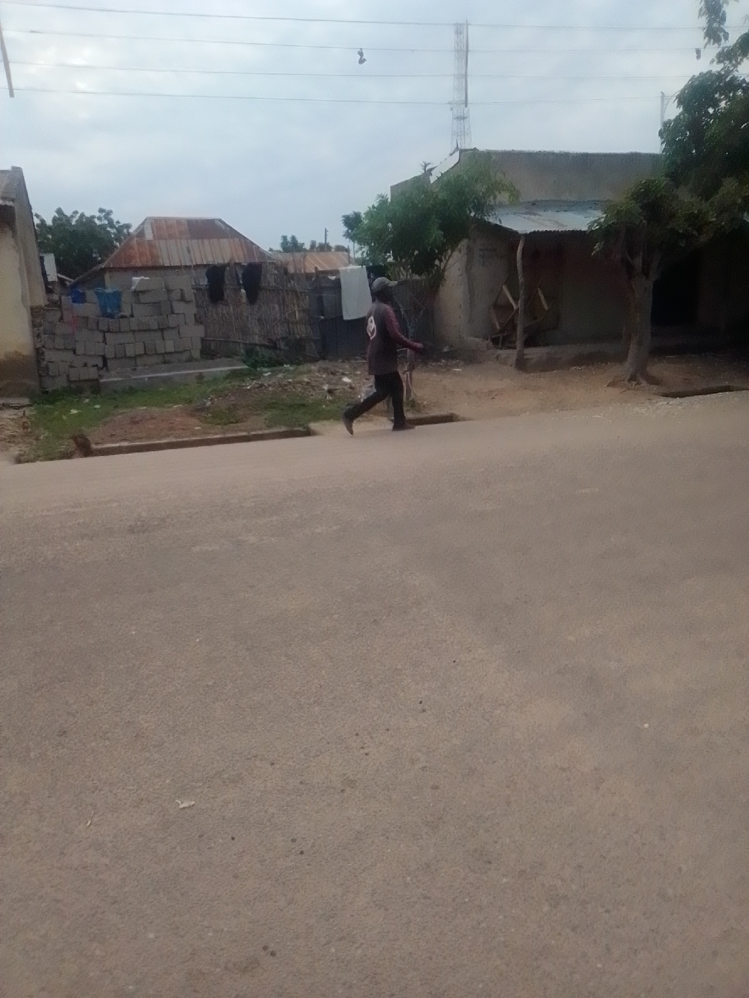 A person is running across a paved road in Liji Hill, Nigeria. The person, wearing a dark shirt and dark pants, is in motion with their left leg extended forward. In the background, there are several buildings with corrugated metal roofs and some with walls made of stacked bricks. Trees and vegetation are visible around the buildings, and a radio tower stands tall in the distance. The sky is overcast.