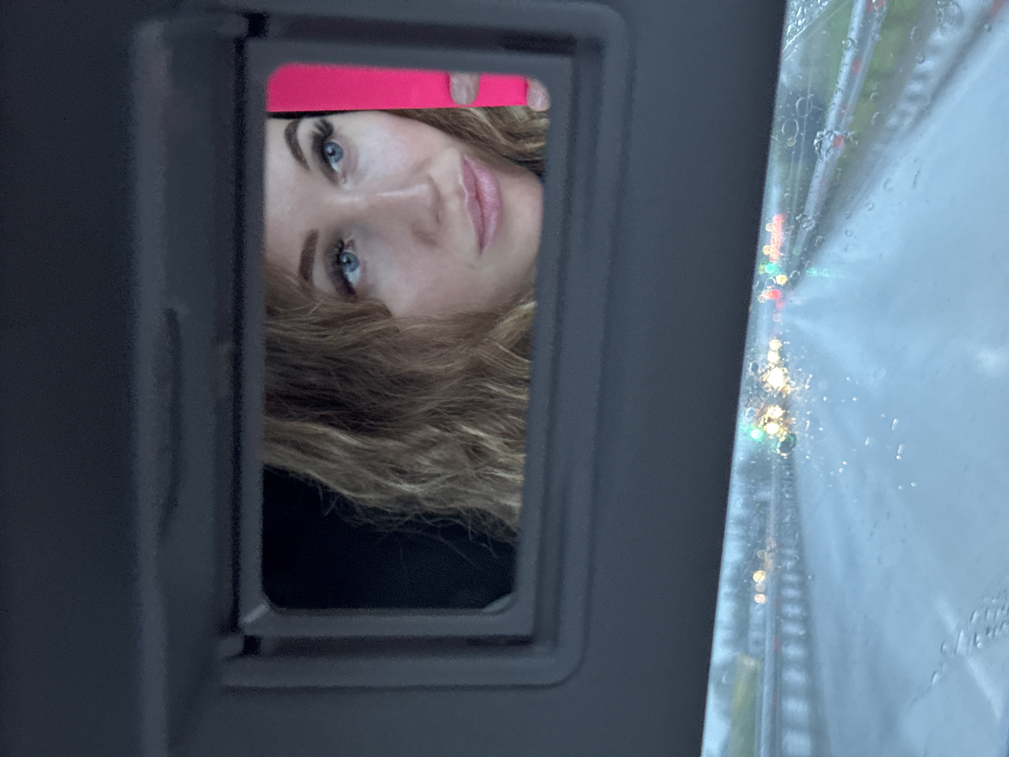 A woman's face is visible in the vanity mirror of a car's sun visor. She is holding a pink object above her head, partially obscuring her forehead. The environment outside the car window appears wet, possibly from rain, and there are blurred lights visible, indicating a road or city environment. It is not possible to determine if this image was taken in Kitsjanovo, Russia.