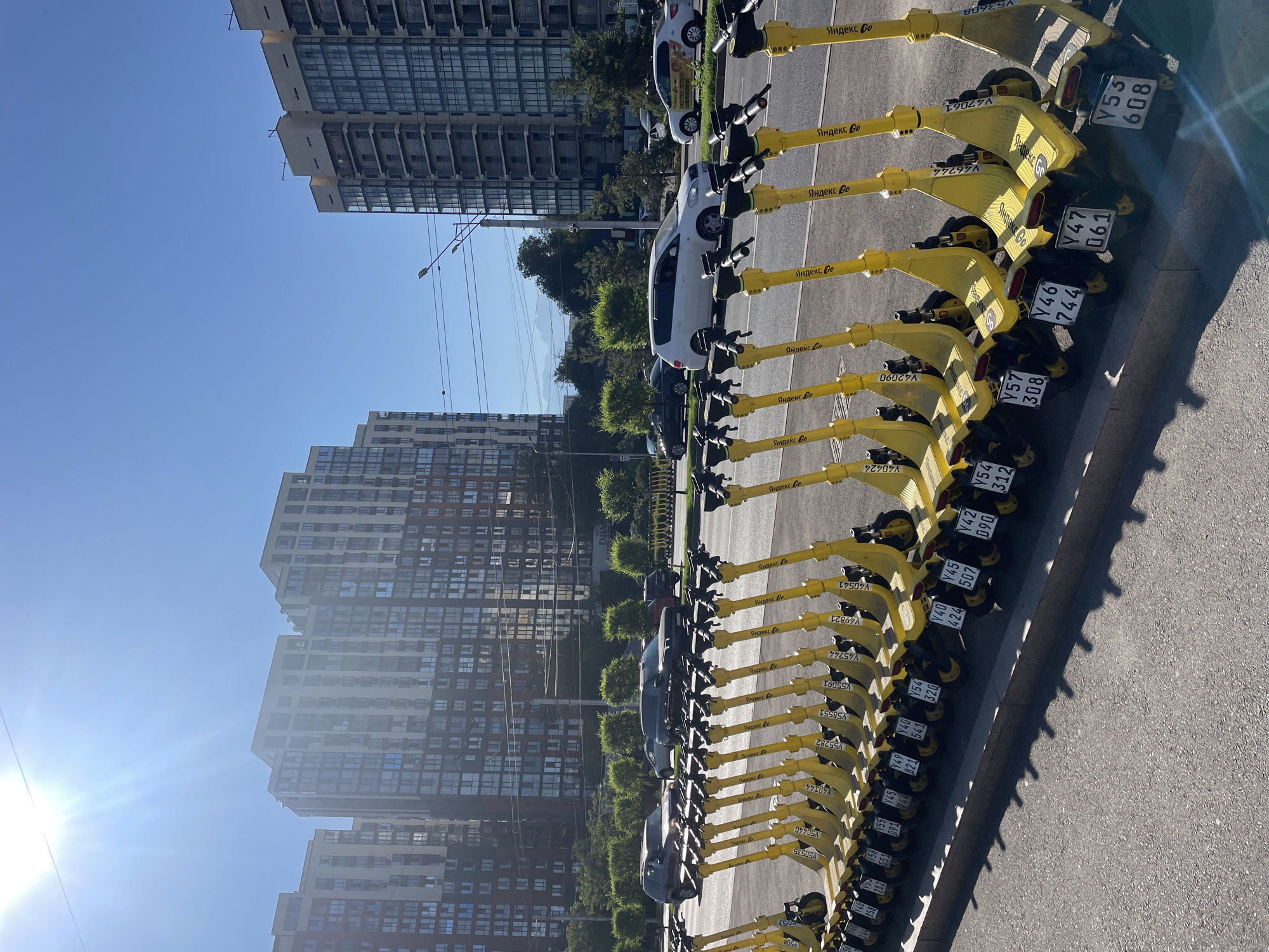 The image depicts a street scene in Almaty, Kazakhstan, on a sunny day. In the foreground, a row of bright yellow electric scooters is neatly parked along a sidewalk. Each scooter has a license plate with alphanumeric characters. Behind the scooters, a paved road with a lane for vehicles is visible, with several cars driving in it. In the middle ground, trees and bushes line the street, creating a green buffer. Further in the background, modern high-rise apartment buildings dominate the skyline. The sky is clear and blue, with the sun creating lens flare effects. The overall impression is one of an urban environment with a focus on shared mobility services.