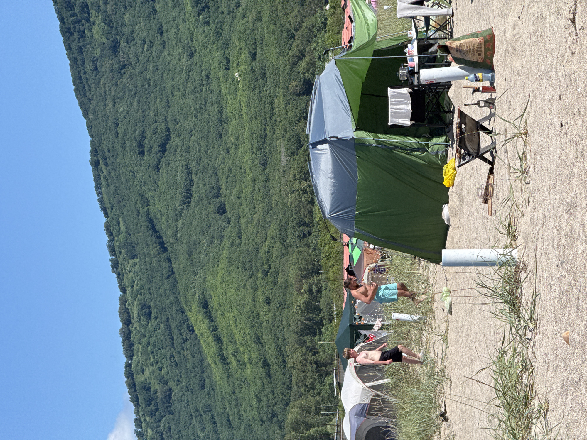 The image depicts a beach campsite with several tents set up on a sandy shore. In the foreground, a large green and grey tent is prominently displayed, with camping equipment such as a portable grill, charcoal bag, propane canister, and a water bottle arranged nearby. Two men are visible walking on the beach between the tents; one is wearing black shorts and the other is in light blue swim trunks. In the background, a densely forested, green mountain range rises sharply, suggesting a coastal or near-coastal location. The sky is clear and blue. The setting is likely a recreational area, possibly a campground on the outskirts of Nakhodka, Russia, given the presence of tents and beach activities, with the mountain providing a natural backdrop.