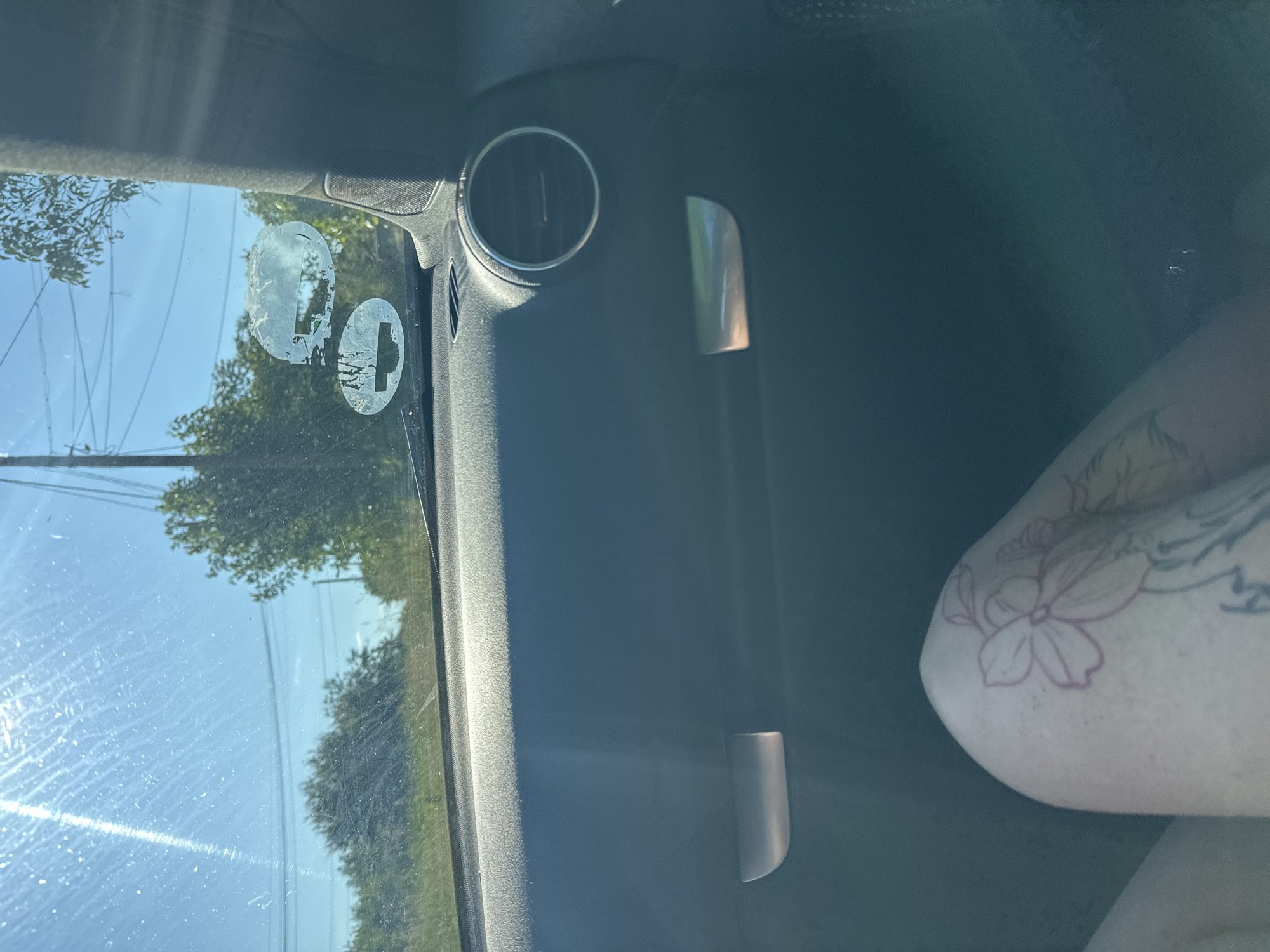 The image captures a sunlit interior view from a car, looking out the passenger side window and across to the vehicle's interior. On the far right, the light-skinned arm and shoulder of a person are visible, featuring a prominent tattoo of a flower, outlined in a reddish-purple hue, along with other grey/black tattoos.
The car's interior, visible on the left and upper parts of the frame, includes a circular air vent in the upper-left quadrant and a metallic, vertically oriented grab handle or trim piece in the mid-right. The side window, appearing somewhat streaky with dust or water marks, showcases bright sun glare. Affixed to the window are two partially peeled, circular stickers; one is mostly residue, while the other clearly depicts a white circle containing a black cross or plus sign symbol.
Through the window, a clear blue sky is visible, along with green trees and foliage suggesting a natural or rural roadside environment. Thin power lines and a utility pole are also discernible against the sky. The bright illumination indicates it is daytime with clear weather. The scene is set in Rogozen, Bulgaria. No specific activity is occurring, and there is no legible text on any visible objects, apart from the cross symbol on the sticker.