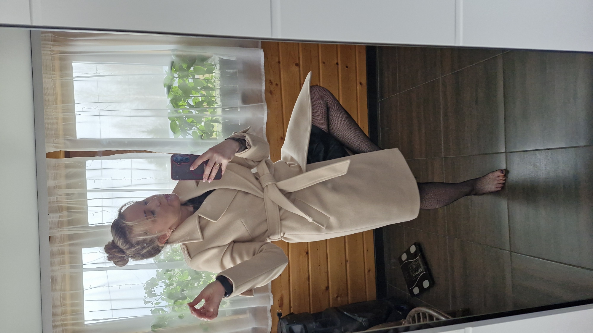 This mirror selfie, taken in Otchor, Russia, depicts a woman posing indoors during daylight hours. She is capturing her reflection using a smartphone with a dark red/brown case, held in her right hand. The woman is dressed in a stylish, long-sleeved, light beige belted coat over a black garment, likely a dress or skirt. She is also wearing black fishnet stockings, revealing her unshod feet. Her hair is styled in a bun, and she wears a ring on her right ring finger and a black smartwatch on her left wrist.
The setting appears to be a room featuring a large mirror that reflects a window draped with white sheer curtains. Beyond the window, green leafy plants suggest an outdoor view. Interior details reflected include horizontal wooden wall paneling and a dark tiled floor. A black bathroom scale, labeled "VITEK" with visible numbers, rests on the floor near her feet, alongside a partially visible metal stool or rack. The image captures a posed moment, showcasing her outfit in a home environment.