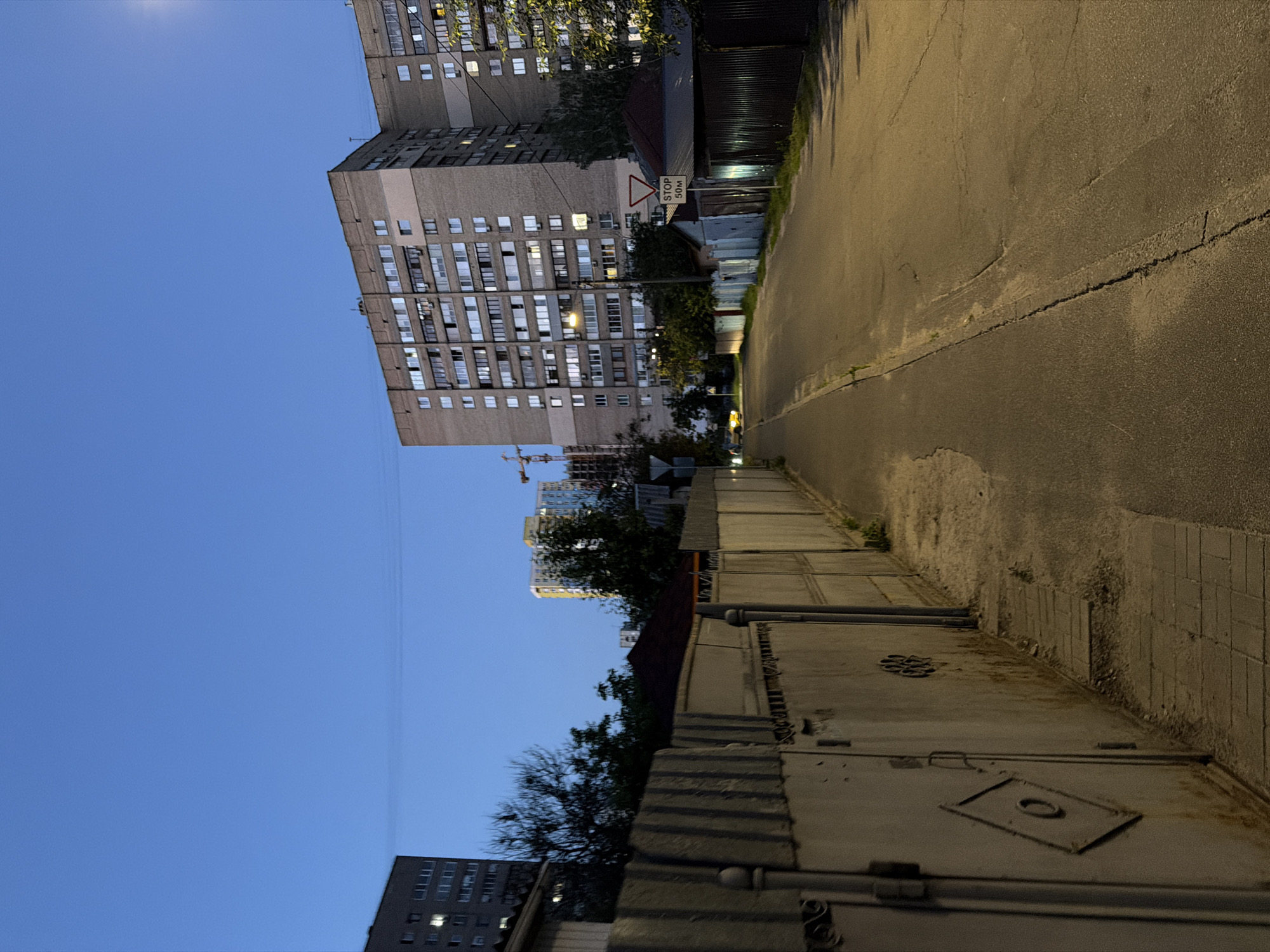 The image depicts a street scene in Kyiv, Ukraine, during twilight. The sky is a deep blue, transitioning from day to night. On the left, a tall apartment building is visible, its windows illuminated. In the foreground, a weathered, corrugated metal fence runs along the side of the street. The street itself is paved with asphalt and shows signs of wear. Further down the road, another, larger apartment building complex dominates the skyline, with many windows lit. A street lamp is illuminated, casting a warm glow. A "STOP 50M" sign is visible on a post. There are no people or vehicles in the immediate scene, though a distant yellow car is present on the road. Construction cranes are visible in the background, suggesting ongoing development in the area. The overall impression is a quiet urban environment in the early evening.