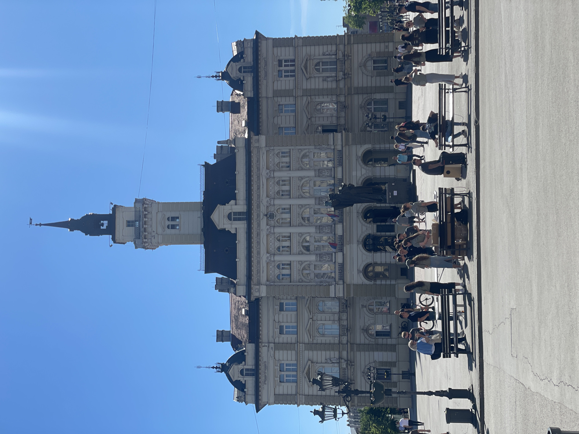 The image shows the City Hall in Novi Sad, Serbia, under a clear blue sky. The building is a multi-story structure with a prominent tower. People are seated on benches in front of the building, some standing nearby. A statue is visible in front of the City Hall. The ground is paved. Street lamps are also visible.