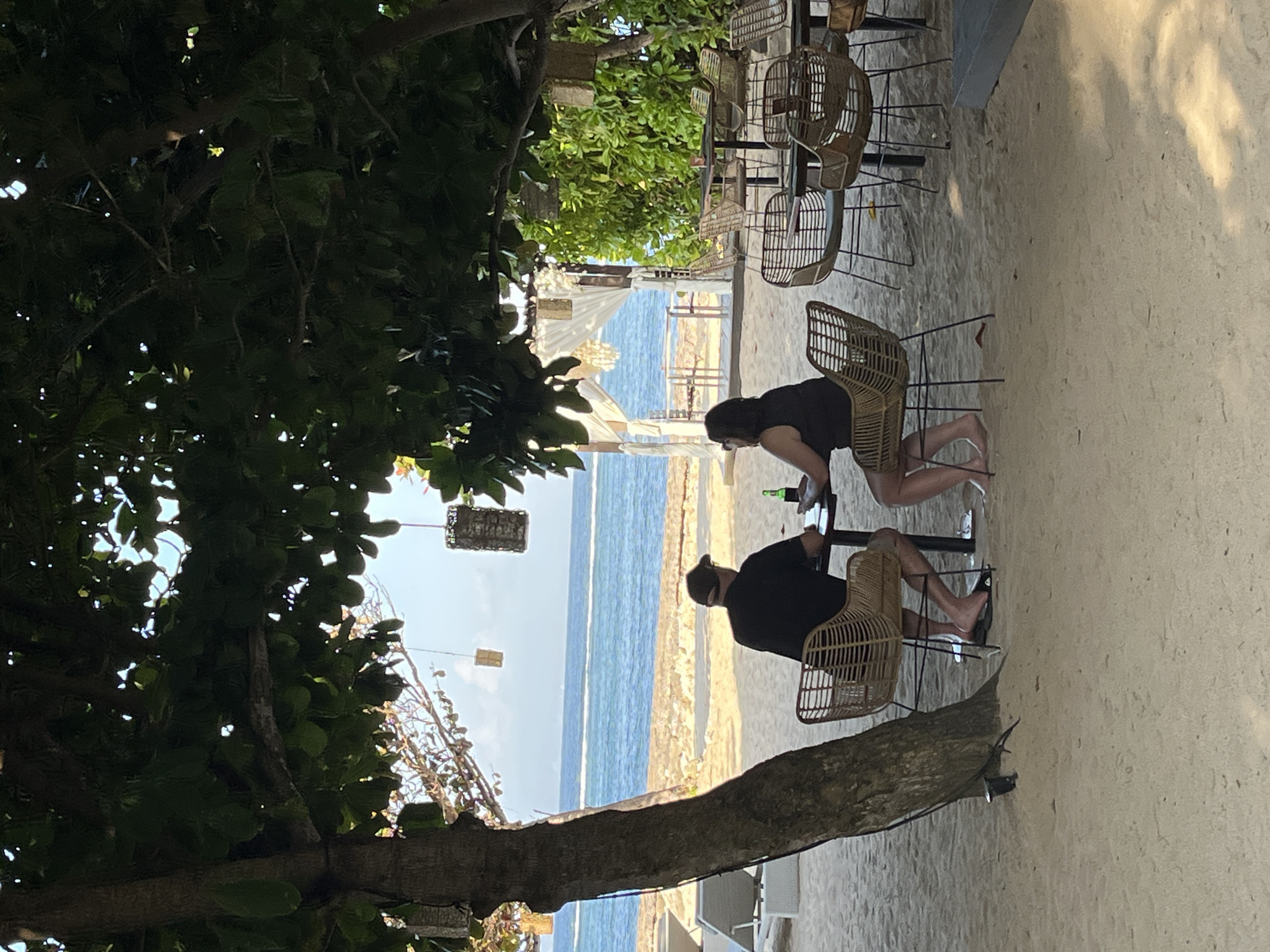 Two individuals, a man wearing a dark t-shirt and a baseball cap, and a woman wearing a dark sleeveless top, are seated at a table on a sandy beach in Kuta Selatan, Indonesia. The man is facing away from the camera, towards the ocean, while the woman is partially turned towards him. They appear to be engaged in conversation. A green bottle is on the table in front of them. They are seated in wicker chairs, characteristic of beachfront establishments.
The setting is a tropical beach with the ocean visible in the background, featuring calm blue water and a clear sky. Lush green foliage frames the right side of the scene, and a large tree with broad leaves provides shade over the dining area. Several other empty tables and chairs are visible, suggesting a restaurant or cafe. In the distance, a white canopy structure with drapes is set up on the beach. The sand is light in color and appears soft. The overall atmosphere suggests a relaxed beachfront dining experience.