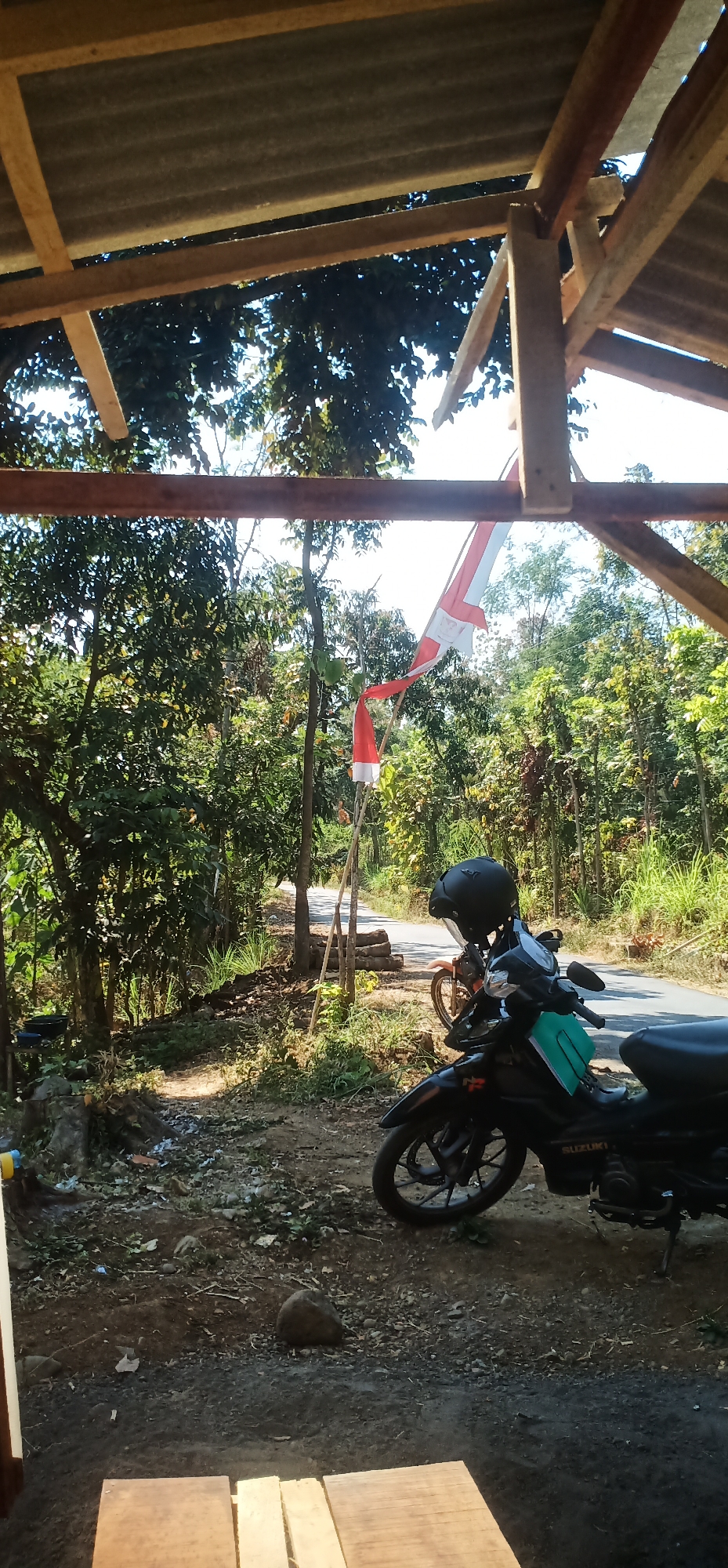 The image captures a roadside scene in Batur, Indonesia, under a partially constructed wooden roof. In the foreground, a black motorcycle with a green accent on its body is parked. A black helmet rests on its handlebars. Behind the motorcycle, another orange motorcycle is partially visible. A paved road recedes into the background, flanked by lush green trees and foliage on both sides. A tall wooden pole with an Indonesian flag, red and white, is planted on the side of the road, with the flag waving in the breeze. The ground is covered with dirt, small stones, and dry leaves. Sunlight filters through the trees, creating dappled shadows. The structure in the foreground appears to be a simple open-sided shelter with a corrugated metal roof supported by wooden beams.