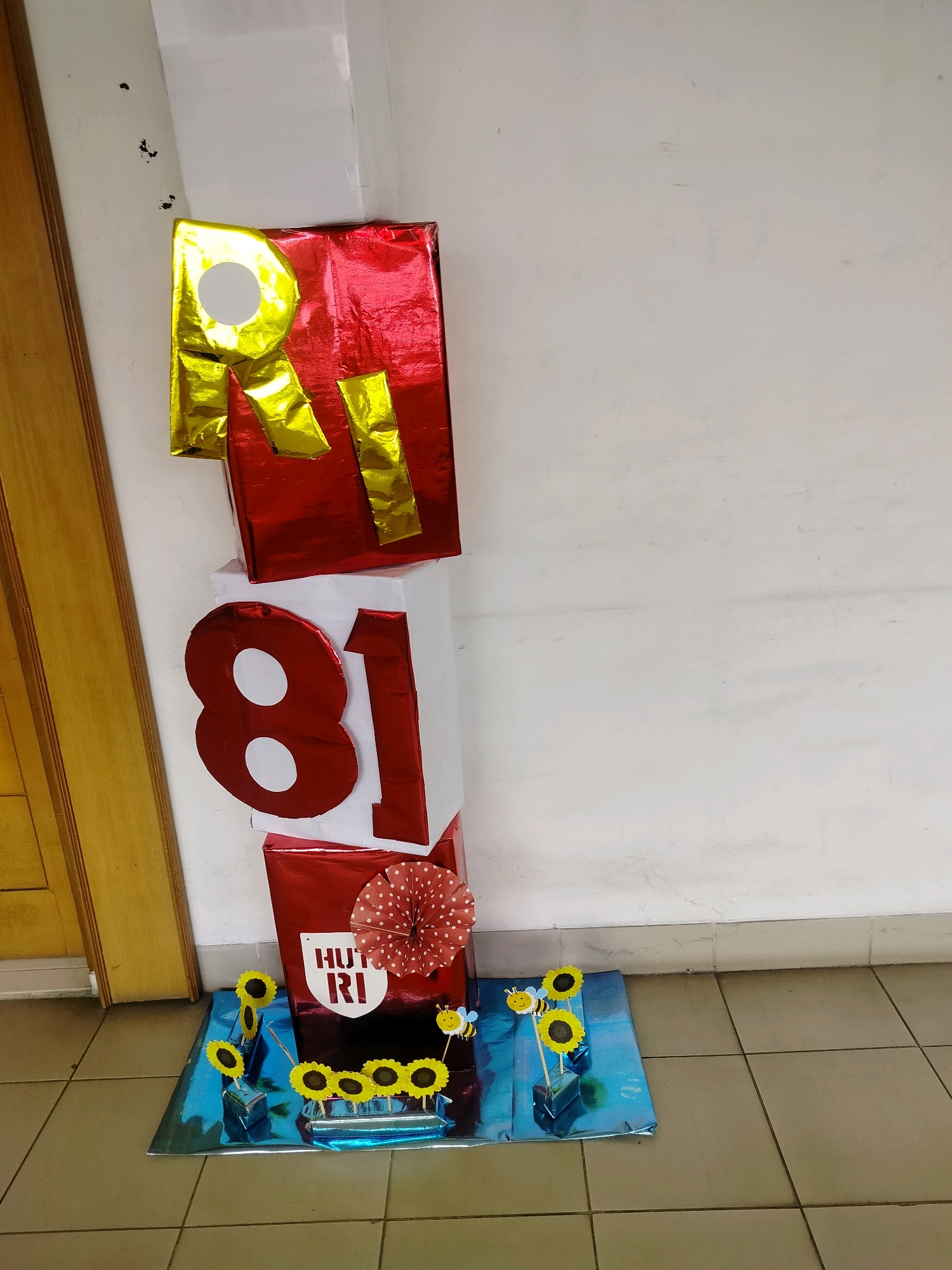 The image shows a stack of three rectangular boxes decorated with wrapping paper and large letter and number cutouts. The top box is wrapped in red foil with gold cutouts forming the letters "RI". The middle box is white with large red cutouts of the number "81" on its side. The bottom box is red and has a white cutout with the text "HUT RI" and a red and white polka dot decorative fan on top of it. In front of the boxes, on a blue metallic mat, are several small sunflower decorations and two cartoon bee figures with smiling faces. To the left of the boxes is a portion of a light brown wooden door frame. The boxes are placed on a tiled floor against a white wall. The scene appears to be an indoor decoration, possibly for a celebration of Indonesia's Independence Day ("Hari Ulang Tahun Republik Indonesia"). The location is specified as East Jakarta, Indonesia.