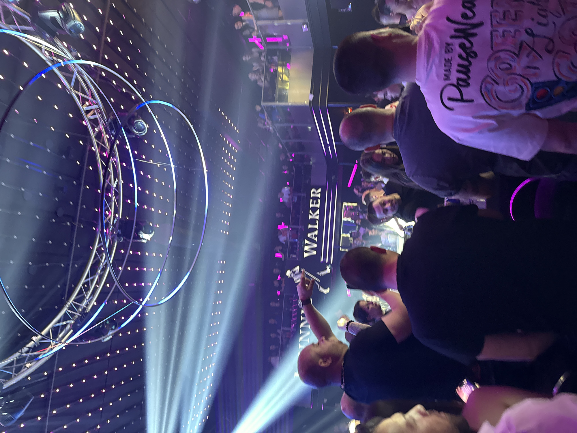 The image shows a crowded indoor venue, potentially a nightclub or concert hall, in Gabrovo, Bulgaria. A large crowd of people is present, many of whom are looking towards a stage or performance area, where there are complex lighting fixtures and a sign that reads "WALKER." A man is holding up his phone to take a photo or video. The lighting is predominantly blue and purple, creating a vibrant atmosphere.