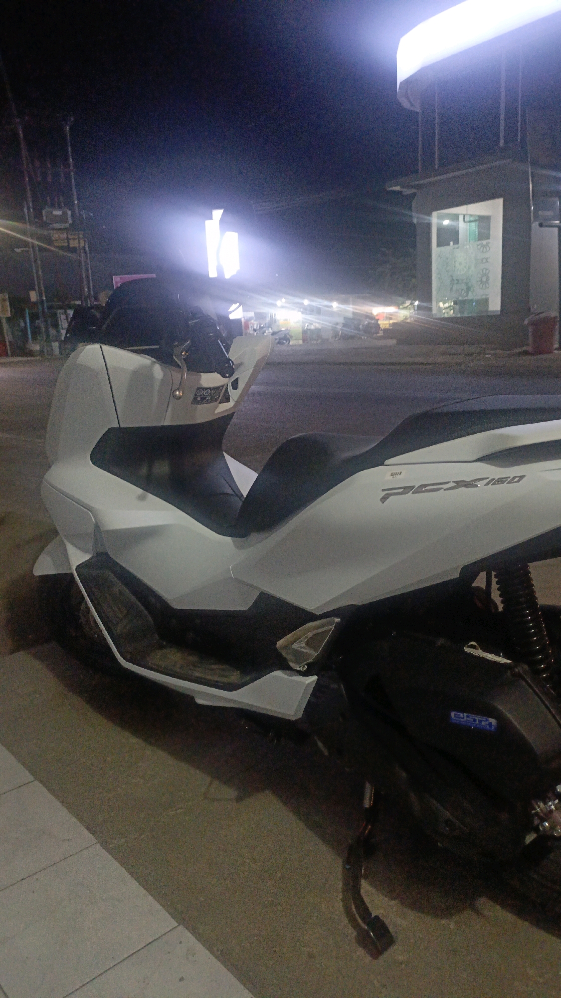 A white Honda PCX 160 scooter is parked on a sidewalk at night. The scooter is facing to the right and is angled slightly towards the viewer. The scooter is clean and appears to be new. The background of the image is blurred and out of focus, but it appears to be a city street with buildings and streetlights. The image was taken in Taliwang, Indonesia.