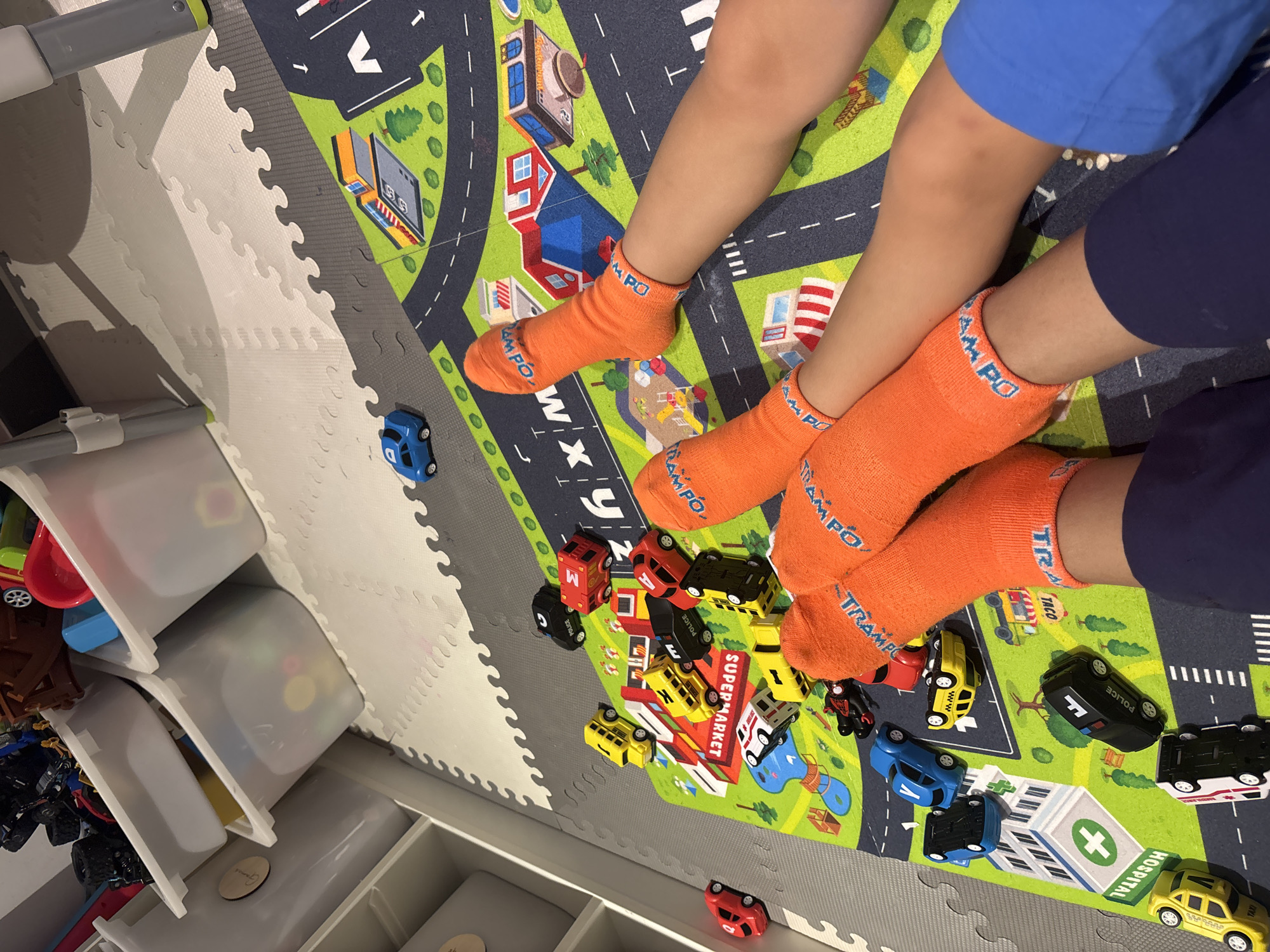 The image depicts a close-up, overhead view of two pairs of children's legs, clad in bright orange socks, resting on a colorful play mat designed to resemble a city road network. Scattered across the mat are various toy vehicles, including cars, police cars, and a taxi, predominantly in red, yellow, and black. A blue toy car is visible in the upper left portion of the image, near some storage bins. The play mat features illustrations of buildings, trees, and a playground. The scene is indoors, likely in a child's playroom, with the textured surface of interlocking foam floor tiles visible beneath the play mat. There is no discernible indication of the location being Dubai, United Arab Emirates.