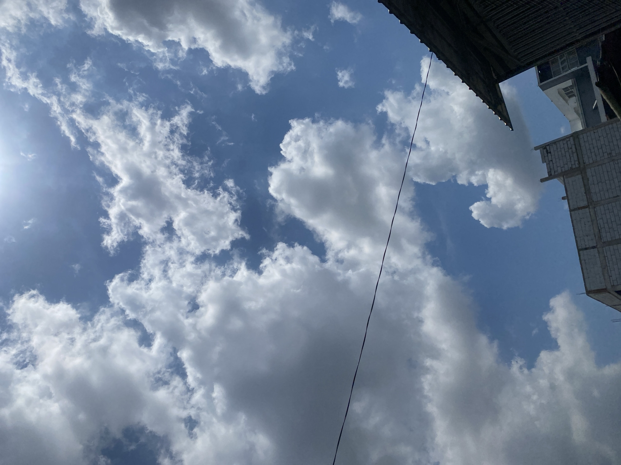 A low-angle shot captures a bright, sunny sky filled with fluffy white cumulus clouds against a vibrant blue backdrop. The sun is visible as a bright white orb in the upper right quadrant of the image, casting a strong light. A thin black wire stretches horizontally across the middle of the frame. In the lower portion of the image, the dark, corrugated metal roof of a building is visible on the right, angled upwards. To the left of the roof, the top section of a white brick building under construction is visible, with exposed rebar at the top. Further to the left and behind the brick building, a modern white and gray multi-story building with windows can be seen. The scene suggests a daytime view from a partially urban or developing area.