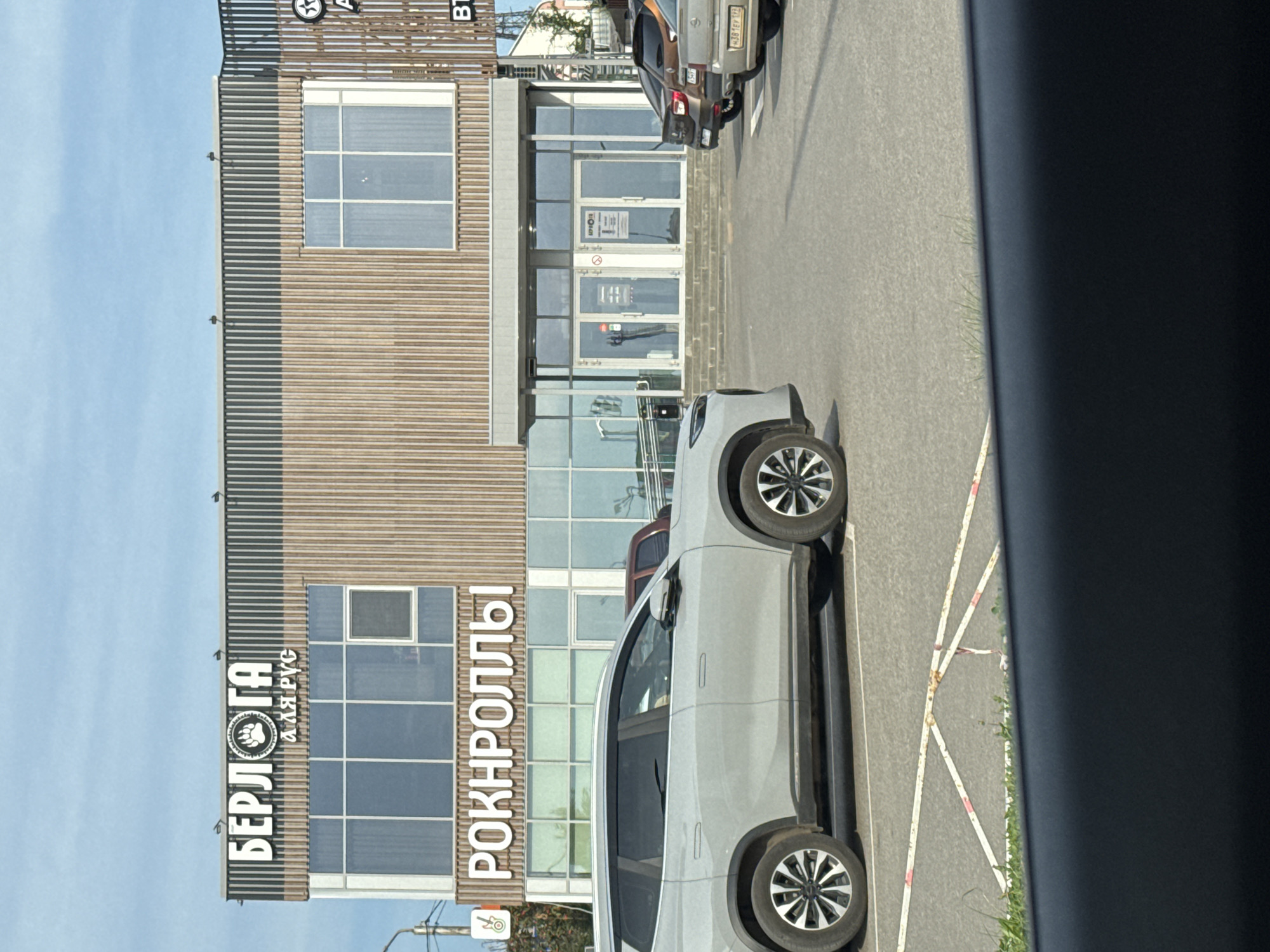 A light gray SUV is parked in a paved lot in front of a modern building. The building has a facade of vertical wooden slats and large windows. Above the windows, a sign reads "БЕРЛОГА" with a paw print logo and below it, "а яя рус". Another sign on the lower part of the building reads "РОКНРОЛЛЫ". The sky is blue with some clouds. Several other vehicles are parked in the lot, and a sidewalk with a ramp is visible near the building entrance. The scene appears to be in a commercial area.