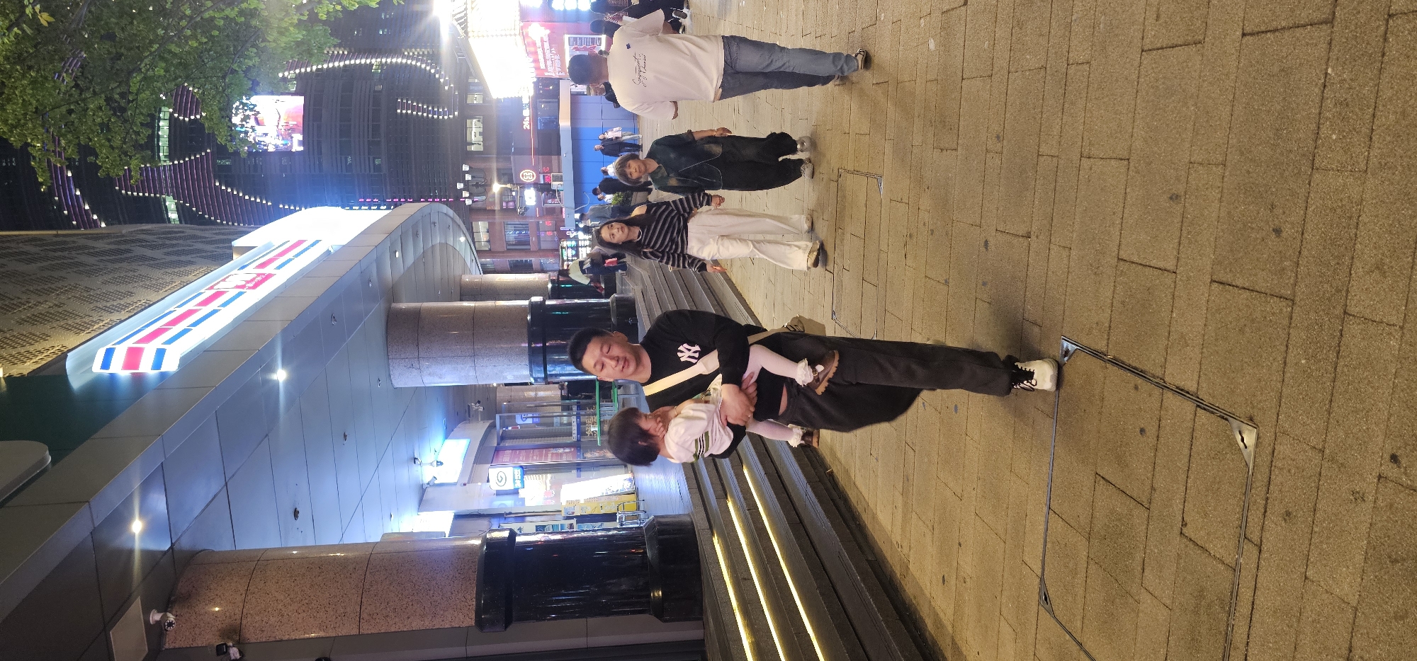 A nighttime street scene in Yuzhong District, China shows a man in a black New York Yankees long-sleeved shirt holding a child in a white dress. They stand on a brick-paved sidewalk adjacent to illuminated steps. Behind them are several people, a building with illuminated signage, and a large modern building with illuminated stripes. The overall lighting is bright.