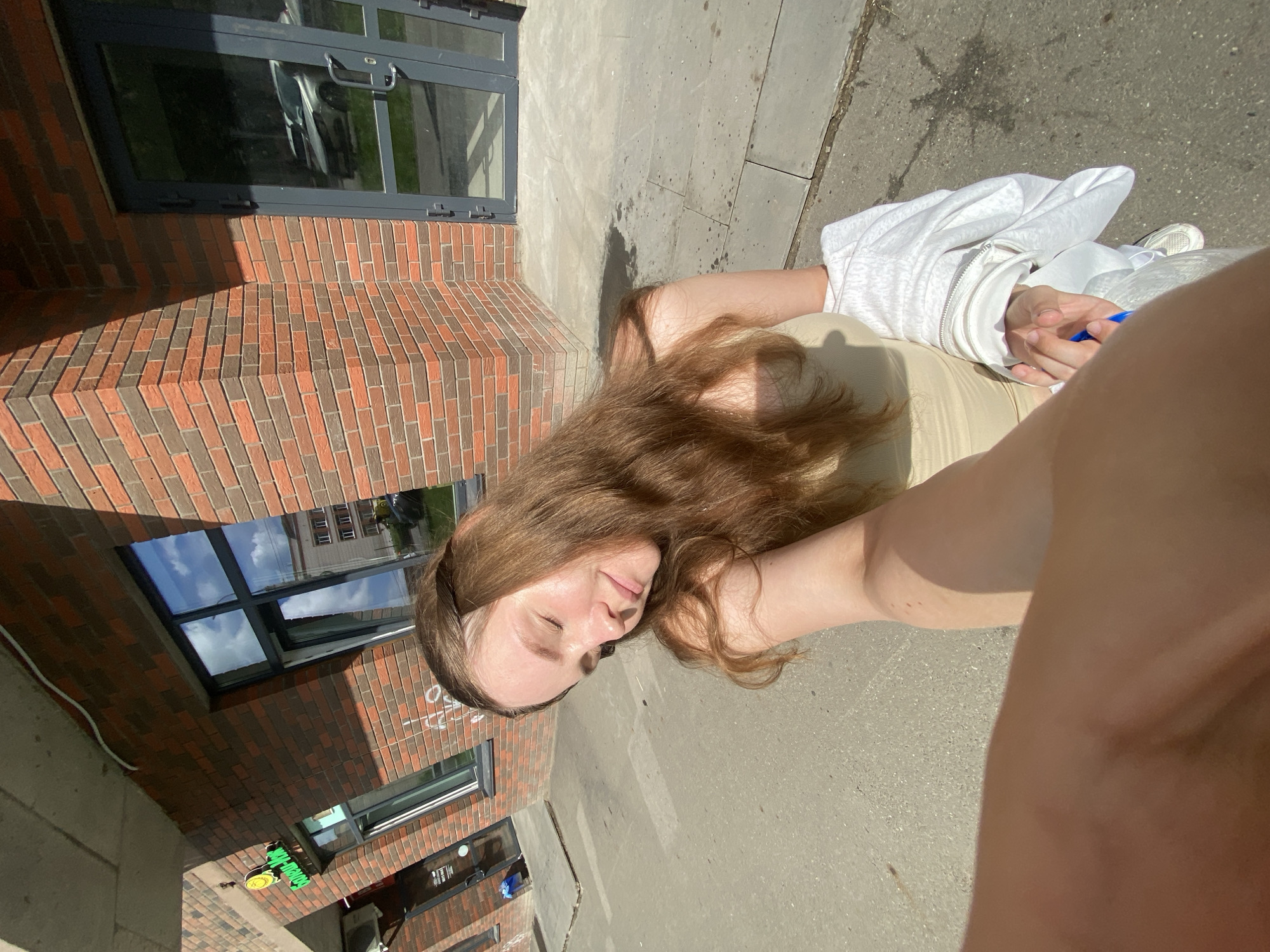 The image, taken outdoors in Izhevsk, Russia, captures a young woman with long, brown hair standing on a paved walkway in front of a brick building. She is wearing a light-colored strapless top and has a white jacket draped over her arm. The woman's eyes are closed, and she appears to be smiling slightly, basking in the sunlight. In the foreground, another person's arm and hand are visible, holding hers. The building features large windows reflecting the sky and other structures. Graffiti can be seen on the brick facade. The overall scene suggests a casual, sunny day in an urban setting.