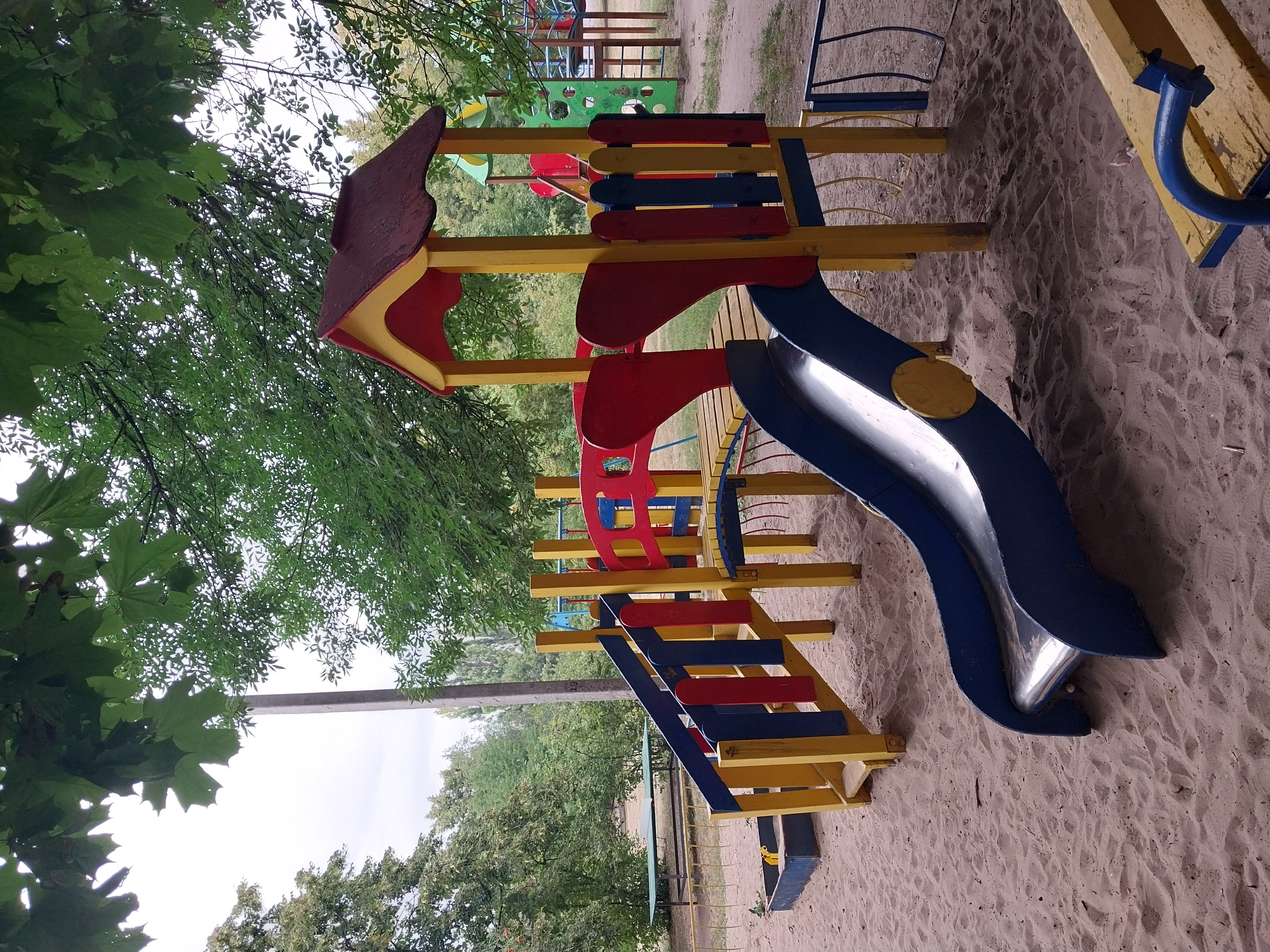 An outdoor children's playground is depicted in Doslidne, Ukraine. The main structure is a multi-colored climbing frame with red, yellow, and blue elements. It features a slide, stairs with protective railings, and a small roofed platform. The ground is covered with sand, and a blue sandbox is visible to the left. In the background, more playground equipment, including a green climbing wall and swings, can be seen. Lush green trees surround the playground, and a utility pole stands to the right of the main structure. The sky is overcast.