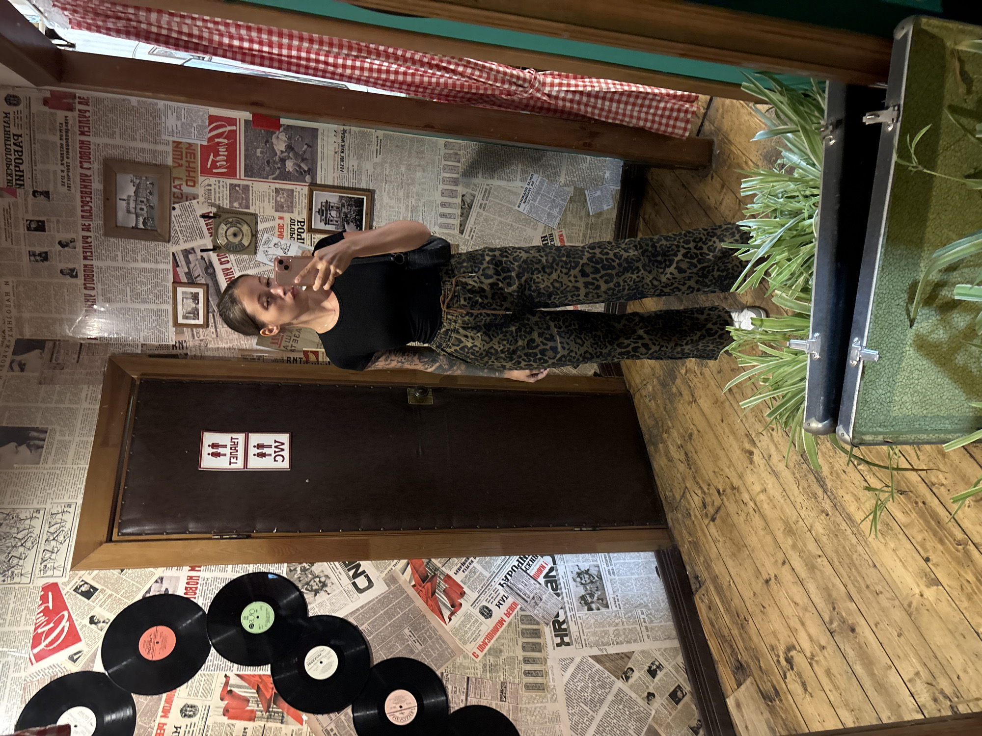 A woman is standing inside of a public restroom with the walls, door, and parts of the floor covered in newspaper clippings and vinyl records. The scene is lit by artificial light.
The woman is fair-skinned, with her brown hair in a low bun, and is wearing a black top and camouflage patterned pants. She is looking at the camera, holding her phone up to take a photo. The door is a dark brown color with a plaque on it that indicates that this is a public restroom.
The walls and floor appear to be made of wood and covered in newspaper clippings and vinyl records. There is a case filled with plants is partially visible in the top-right corner of the image. The location is suggested by the presence of Cyrillic text to be Moscow, Russia.