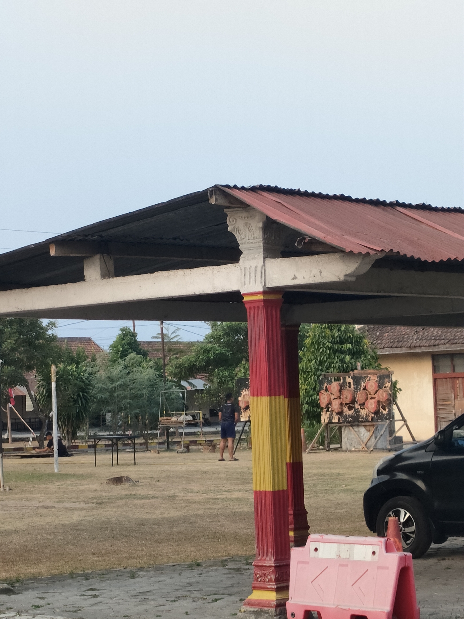 The image captures an outdoor scene in Banguntapan, Indonesia, characterized by dry, light-colored grass covering the ground. In the foreground, a bright pink plastic barrier with white reflective strips is visible. A black car is parked to the right of the image, facing left.
Dominating the mid-ground is a covered structure with a red corrugated metal roof supported by white beams. Two large pillars, painted red and yellow, support the front of this structure. Behind the structure, several trees and buildings with tiled roofs are present. In the background, a target board with circular patterns is mounted on a wooden stand.
The scene appears to be a public area, possibly a park or an open field. One individual, wearing a dark t-shirt and blue shorts, is standing in the mid-ground. Another individual, dressed in dark clothing, is sitting on the ground in the left mid-ground. The overall atmosphere suggests a warm climate with dry conditions.