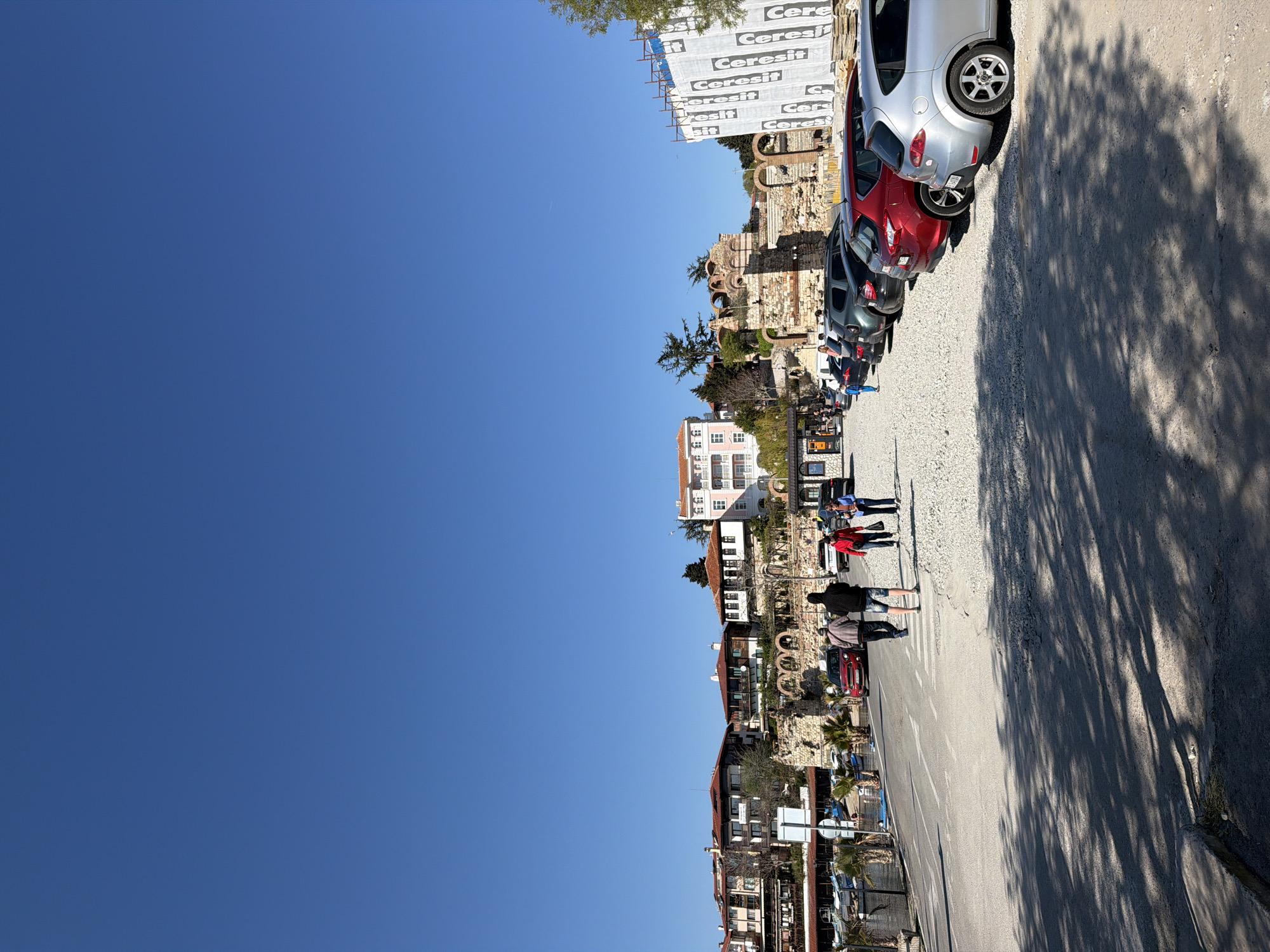 The image depicts a street scene in Nesebar, Bulgaria. The perspective is rotated 90 degrees clockwise. A line of parked cars, including silver and red models, runs along the right side of the frame, creating a boundary along the street. Shadows from trees are cast across the pavement.
On the opposite side of the street, there are buildings, including residential structures and what appears to be the remnants of ancient architecture. A building covered with a construction banner is visible behind the ruins. Several people are seen walking along the sidewalk. Overhead, a clear blue sky dominates the upper portion of the image.
