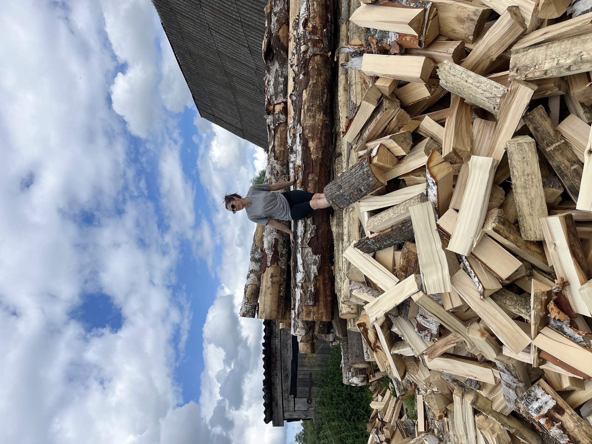 A person is sitting on a stack of large logs in front of a pile of cut firewood. The person, wearing a gray t-shirt and black shorts, has their legs crossed and is looking to the left. Behind the logs, a rustic wooden shed is visible to the left, and a building with a dark, corrugated roof is to the right. The scene is set outdoors under a cloudy blue sky. The location is Уральское, Russia.