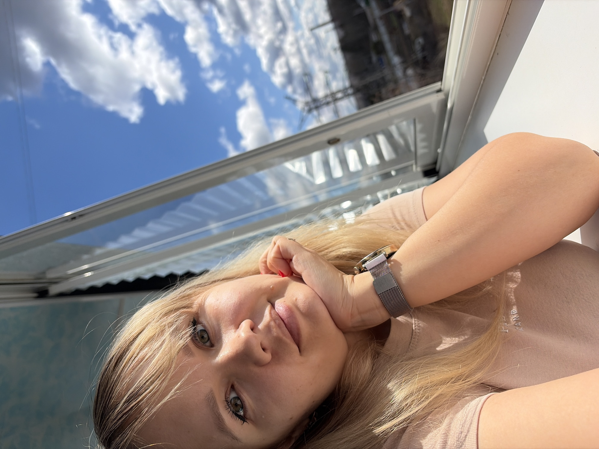 A bright, upward-angled portrait shot captures a young woman with fair skin, blonde hair, and striking green eyes, looking directly at the camera. She appears to be lying down or reclining, with her head slightly tilted. Her blonde hair cascades around her face and shoulders. Her lips are slightly parted, and her expression is calm and relaxed.
She is wearing a light beige or pale pink long-sleeved top, and her left arm is bent, with her hand resting gently near her chin. A silver-toned watch with a mesh strap is visible on her left wrist. A hint of red nail polish can be seen on one of her fingers.
The background primarily features a clear blue sky with fluffy white clouds, visible through what appears to be a large window or skylight directly above her. The window frame, made of light-colored material, runs horizontally across the top portion of the image (as if rotated). Beyond the window, on the right side, blurred dark structures, possibly power lines or distant buildings, are faintly visible, suggesting an outdoor or semi-outdoor setting. The bright illumination indicates it is daytime, likely sunny.
On her top, a faint, light-colored embroidered or printed text is visible, which appears to contain the letters "AND" or "LLAND" but is largely illegible.