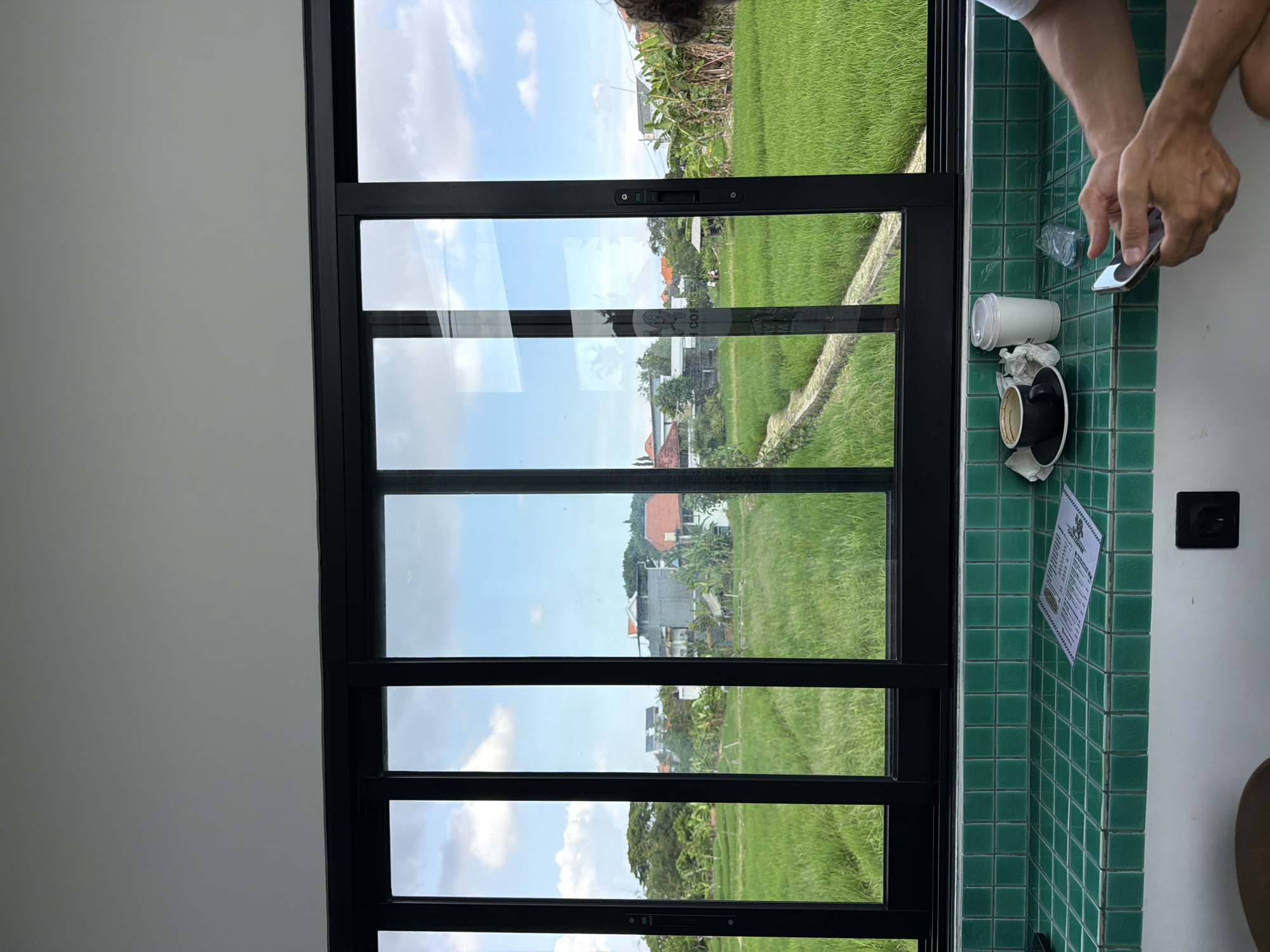 The image shows an interior view through a large, black-framed window, offering a direct view of a vibrant green rice field and scattered buildings, consistent with the landscape in North Kuta, Indonesia. The window has multiple panes, creating a grid-like division of the outdoor scenery. To the right of the window, there is a tiled green wall. On the tiled surface, there are discarded coffee cups, napkins, a plastic water bottle, a menu, and an electrical outlet. A person's hands and arms are visible as they are interacting with a mobile phone.