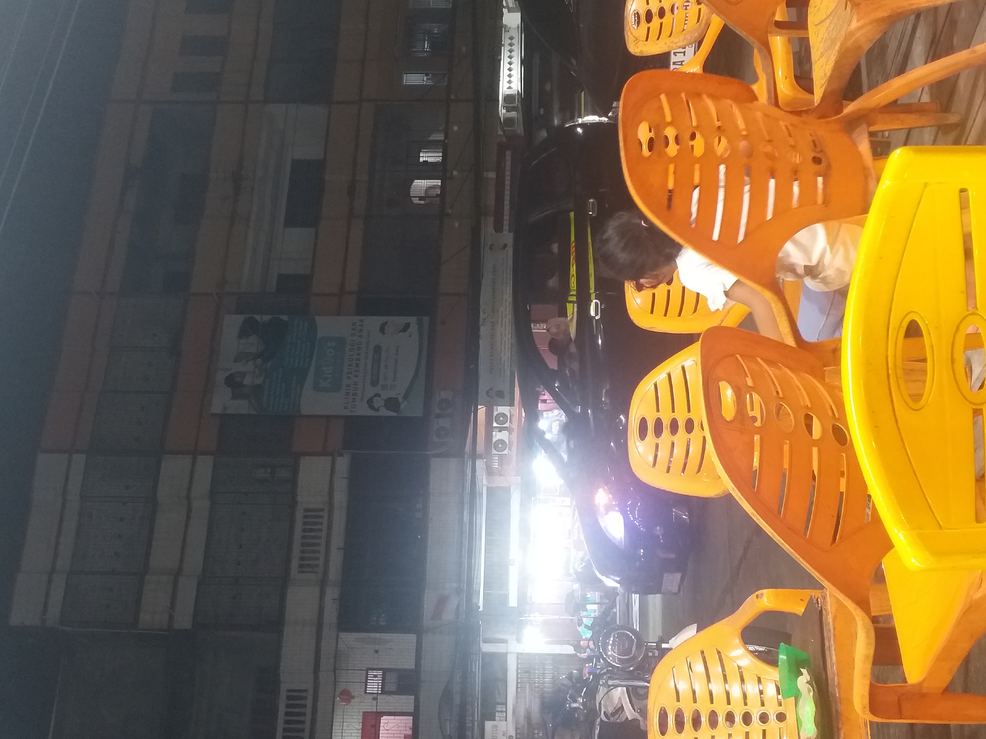 A night-time, eye-level, outdoor medium shot captures a scene outside a building in Dumai City, Indonesia. In the foreground, the back of a child wearing a white t-shirt is visible, seated in an orange plastic chair. Several other orange and yellow plastic chairs are arranged around the area. In the mid-ground, a dark-colored car is parked, with its headlights illuminated. Behind the car, a building with multiple stories is visible, featuring windows and signage. The lighting suggests an urban environment at night, possibly a commercial or residential area.
