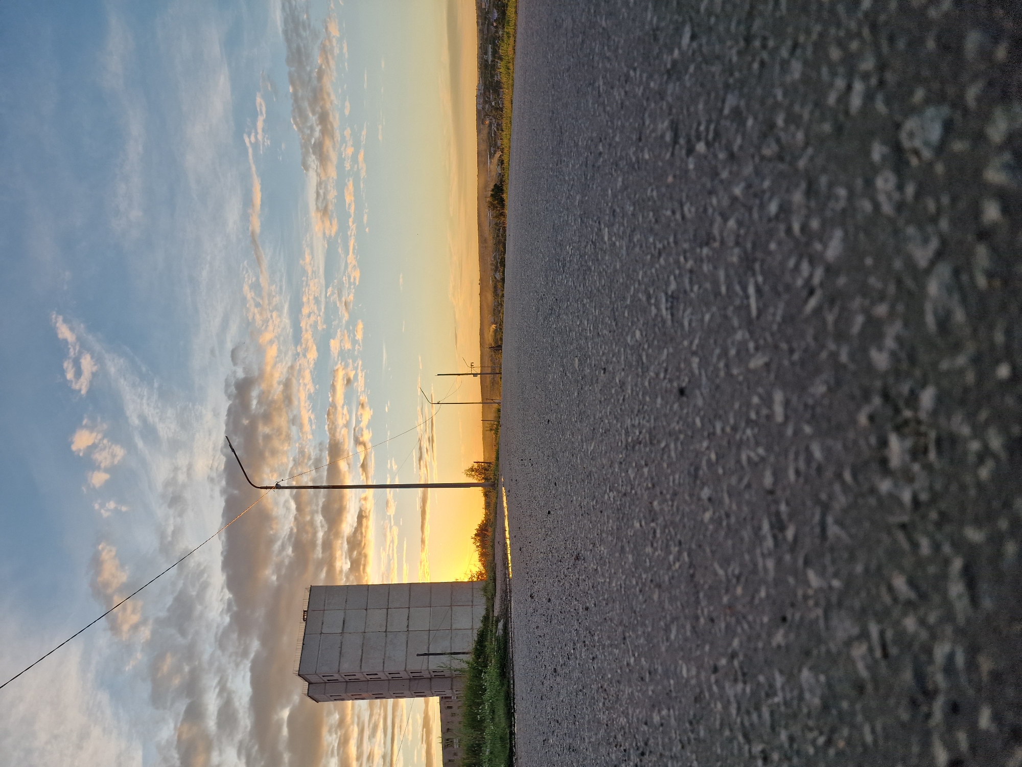 A low-angle shot shows an asphalt road in the foreground, with a tall, grey, multi-story building to the left. A utility pole with a light fixture stands near the building, and another pole is visible further down the road. The sky above is a mix of blue and scattered clouds, with the sun setting on the horizon, casting an orange glow. In the distance, a town or city is visible, with houses and trees. The scene appears to be outdoors in an urban or suburban environment.