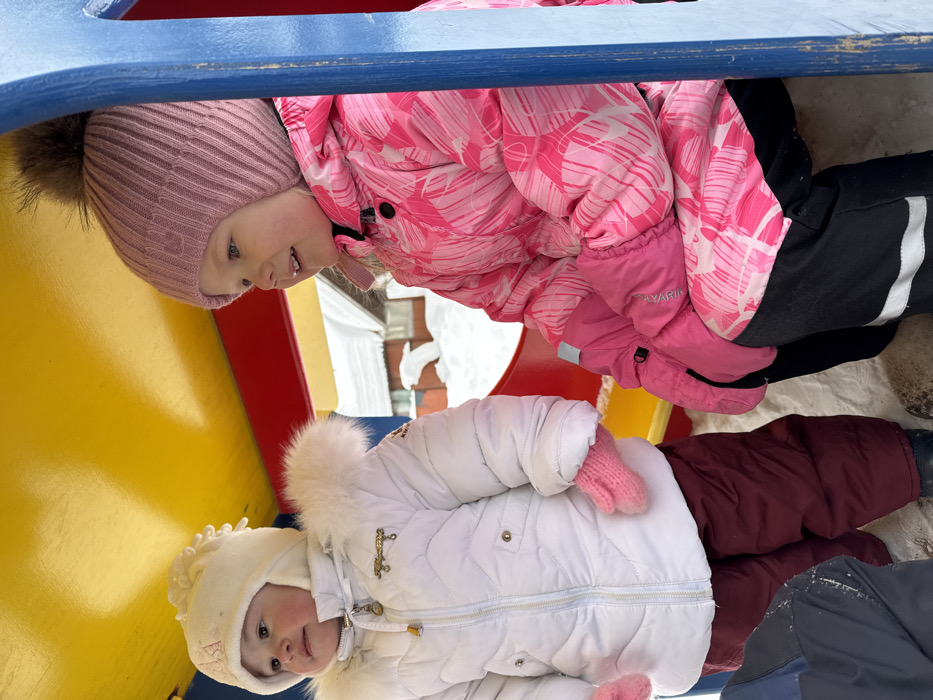 Two young children are observed within a brightly colored outdoor playground structure in Zaosinovo, Russia, during what appears to be a snowy winter day. The structure features a yellow ceiling, red side panels, and blue vertical supports.
The child on the left, a younger individual, stands facing slightly left, looking upward with a serene expression. This child wears a white quilted winter coat with a fur-trimmed hood, a white knitted hat with ear flaps, and pink knitted mittens. Dark red snow pants are visible below the coat, and dark footwear is partially obscured by snow on the ground.
The child on the right, an older individual, is seated and positioned towards the right, looking out of frame with a slight smile and open mouth. This child wears a pink winter jacket with a white abstract pattern, a ribbed pink knitted hat topped with a large, fluffy brown faux fur pom-pom, and bright pink winter gloves. Black snow pants with white reflective stripes near the cuffs complete the attire.
The ground within the structure and in the background is covered with snow. Beyond the structure, a building with a white, snow-covered roof is partially visible, indicating a potentially residential or rural setting. The lighting is bright and diffused, consistent with an overcast winter environment.