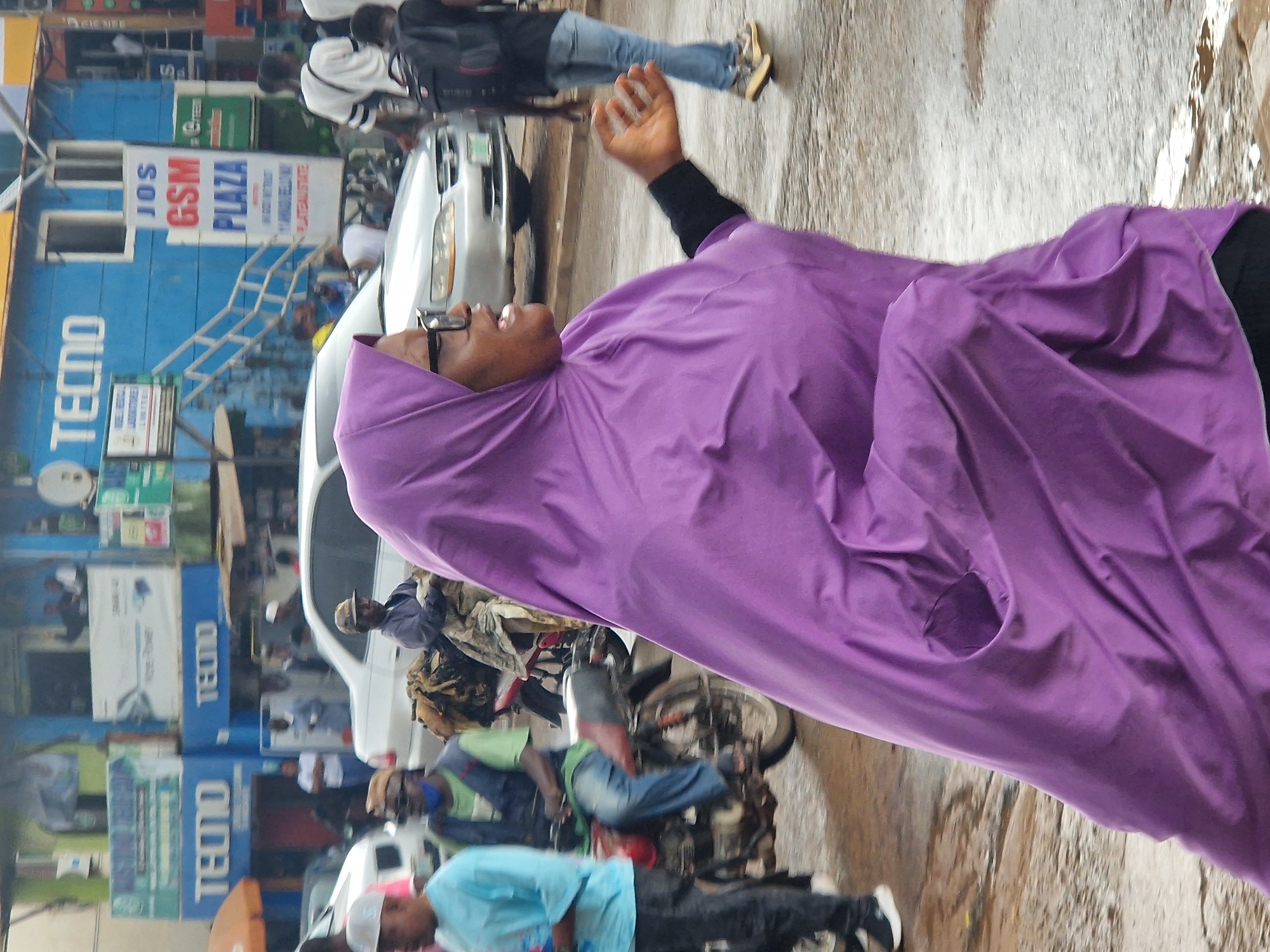 The image captures a street scene in Jos, Nigeria. In the foreground, a woman wearing a vibrant purple hijab and glasses is walking towards the right, with her mouth open as if speaking or reacting. She has a black sleeve on her left arm. Behind her, a silver sedan car is visible. To the left of the woman, a man in a blue t-shirt and dark pants is standing, looking towards the right. Further left, another man on a motorcycle is wearing a green shirt, a dark vest, and blue jeans, with a cap on his head. The motorcycle has some items strapped to its rear. In the background, a blue building with "TECNO" signage is prominent, suggesting a commercial area. Other buildings and signs, including "JOS GSM PLAZA", are also visible. The ground appears wet, indicating recent rain. People are scattered throughout the scene, some walking and others on motorcycles. The overall impression is that of a busy urban street environment.