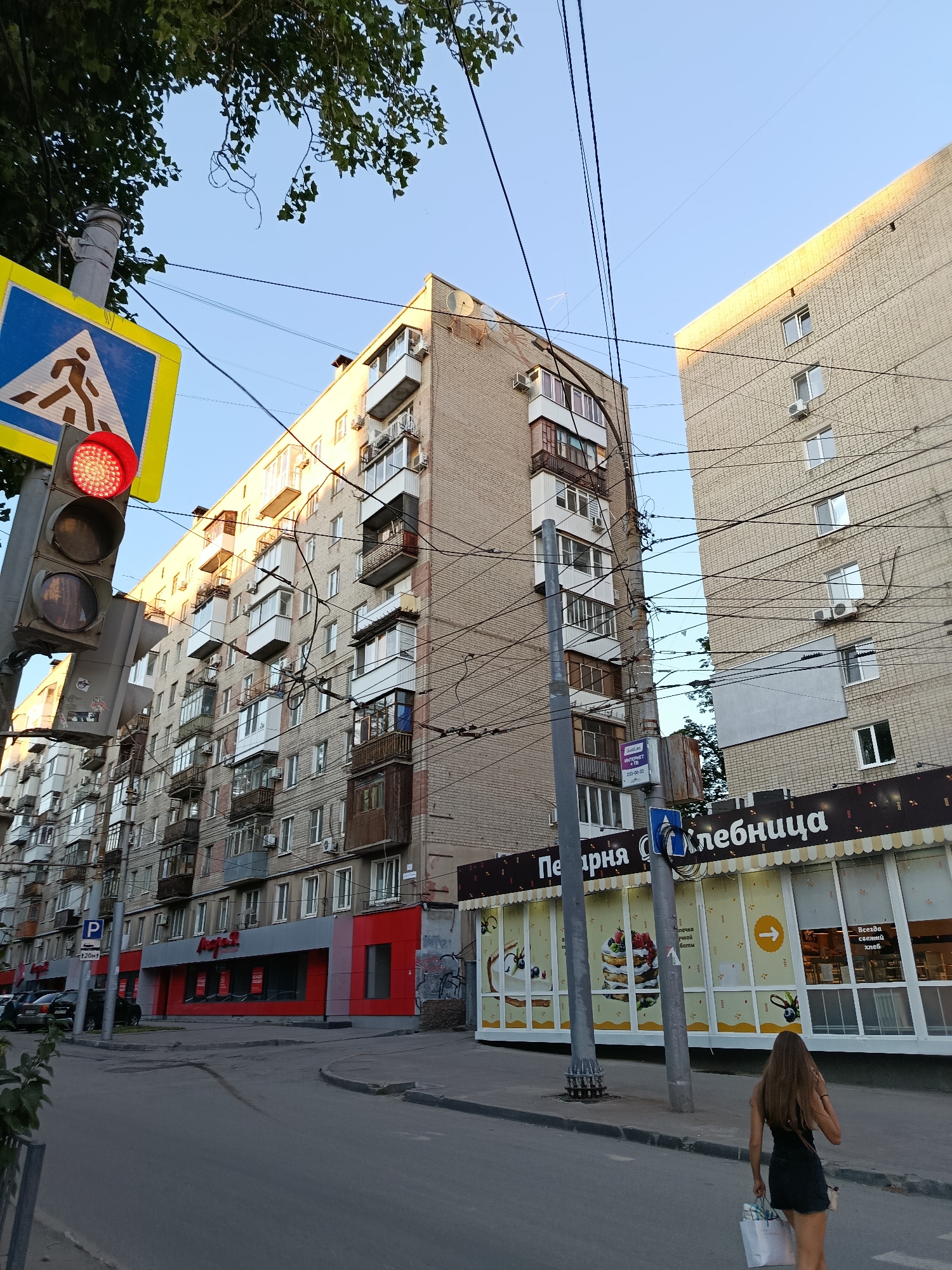 This is a street-level, eye-level, wide-angle photograph taken in Saratov, Russia. The scene captures a street with a sidewalk and two multi-story brick buildings. On the left is a pedestrian crossing sign with a traffic light showing a red signal. The building on the left has a red and gray facade on the ground floor, with several stories of apartments above, many with balconies. The building on the right is lighter-colored brick and also has multiple stories of apartments. A bakery with large display windows is visible on the ground floor of the building on the right, featuring images of cakes and pastries and Cyrillic text that reads "Пекарня Хлебница" (Bakery Breadbox). Numerous electrical wires crisscross the sky between the buildings. A young woman with long brown hair is walking away from the camera on the sidewalk in front of the bakery, carrying shopping bags. The sky is clear and blue.
