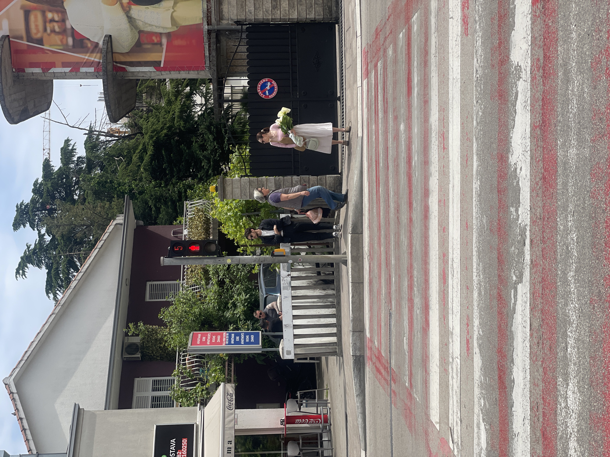 A woman is standing in a crosswalk holding a bouquet of white flowers, while three other people are standing on the sidewalk. A pedestrian crossing signal is visible, displaying a red light. A Coca-Cola branded cafe is on the corner. A building advertising apartments, rooms, and "zimmer" is on the left side of the image. The location is Budva, Montenegro, based on the signage referencing "VILLA MIRAY BUDVA." The crosswalk is marked with alternating red and white stripes. A black gate is visible behind the woman.