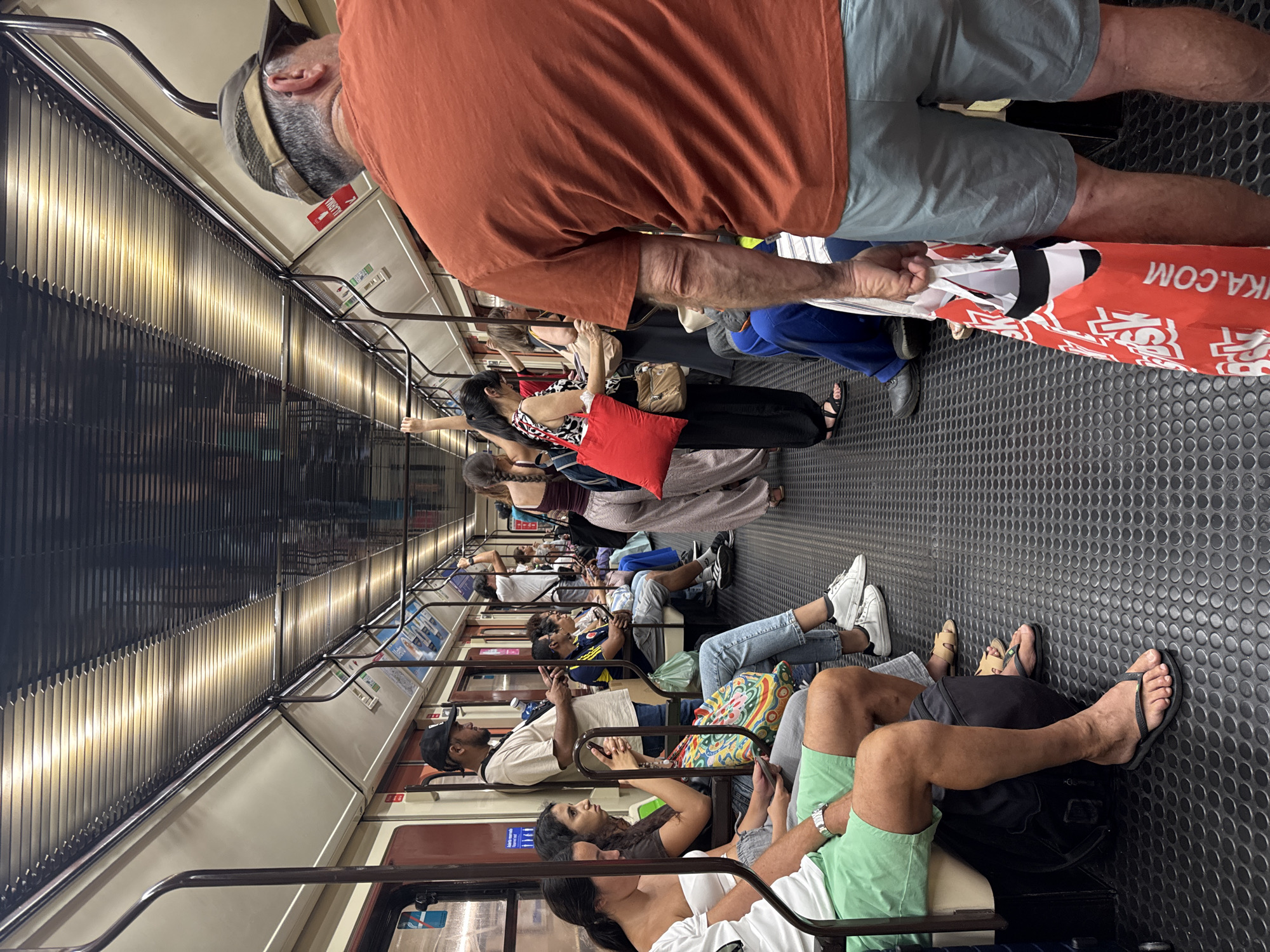 The image depicts the interior of a crowded Metro de Madrid train car. Passengers are standing and seated, occupying most of the available space. The train car features the characteristic red and cream color scheme, with metal poles for support and ribbed flooring. The ceiling has a distinctive striped pattern with embedded lighting. Several individuals are visible in various states of engagement, some looking at their phones, others conversing, and some simply observing their surroundings. One person in the foreground on the right is wearing an orange t-shirt and grey shorts, carrying a red and white reusable shopping bag. On the left, a person in green shorts and a white t-shirt is seated. The overall atmosphere suggests a typical commute on public transportation in Madrid.
