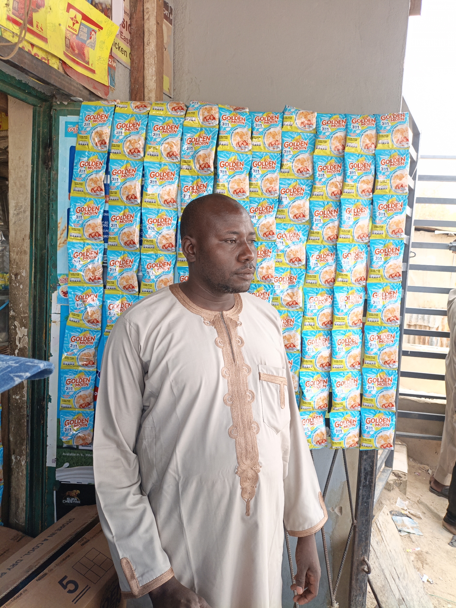The image depicts a man standing in front of a display of "Golden Morn 3 in 1" cereal packets in what appears to be a shop or stall in Kawaji, Nigeria. The man is dark-skinned and bald, wearing a light-colored traditional Nigerian garment with brown embroidered patterns on the front.
Behind him, a wall of Golden Morn cereal packets is visible. There are several rows and columns of the blue and yellow packets creating a grid-like pattern. To the left is the storefront with other food advertising.
To the right of the wall of cereal is an open outdoor space with metal bars. Another person is partially visible standing by metal bars. Boxes are visible in the bottom left corner.