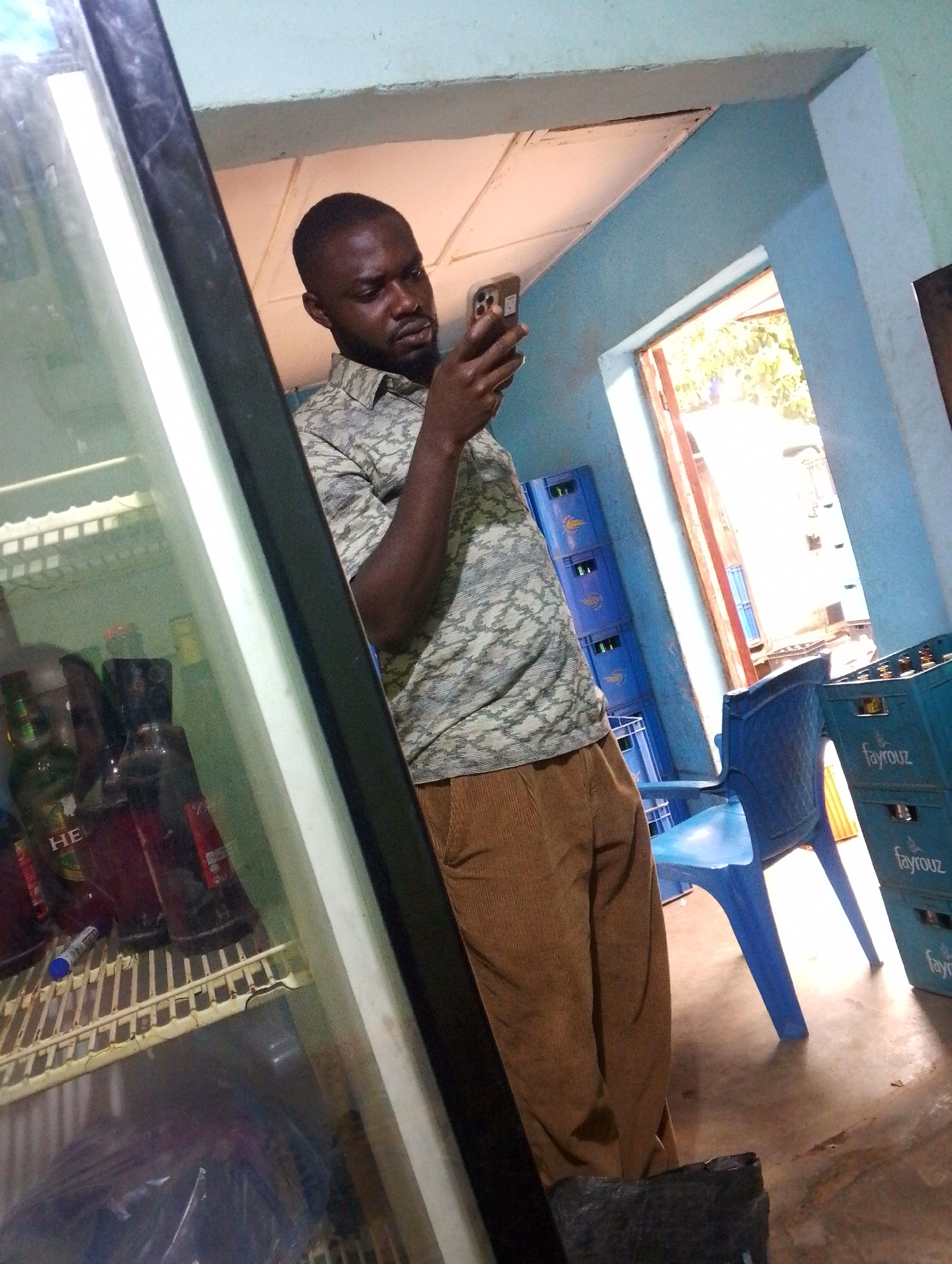 A man with short hair and a beard is standing in what appears to be a small convenience store or bar in Unknown, Unknown. He is holding a mobile phone and looking at it intently. The man is wearing a patterned shirt and brown corduroy pants. Behind him is a doorway leading to the outside. To his left is a refrigeration unit filled with various bottles and cans, presumably beverages. In the background, there are blue plastic crates stacked on top of each other, and a blue plastic chair is visible. The walls of the room are painted in a light blue color. The lighting inside seems to be a mixture of natural light coming from the open doorway and artificial light from inside the establishment.