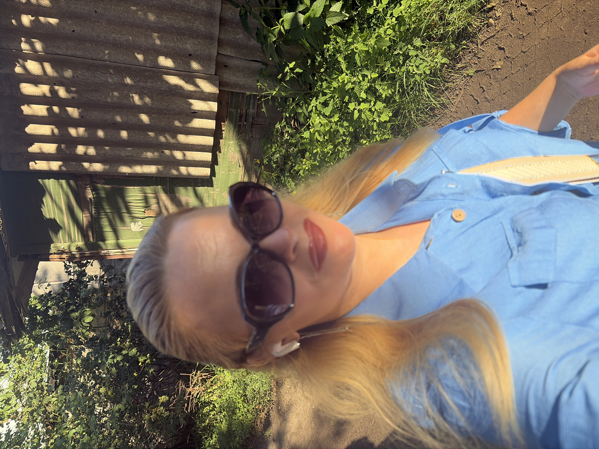 A blonde woman with sunglasses and a blue shirt is standing outdoors in what appears to be a rural or neglected area, possibly near an old building. The building has a weathered exterior with green paint and corrugated roofing. Sunlight casts shadows on the building's surface. Green foliage and a dirt path are visible in the foreground and surrounding the woman. She is wearing white earbuds and has red lipstick on. The woman is holding a camera, likely taking a selfie.