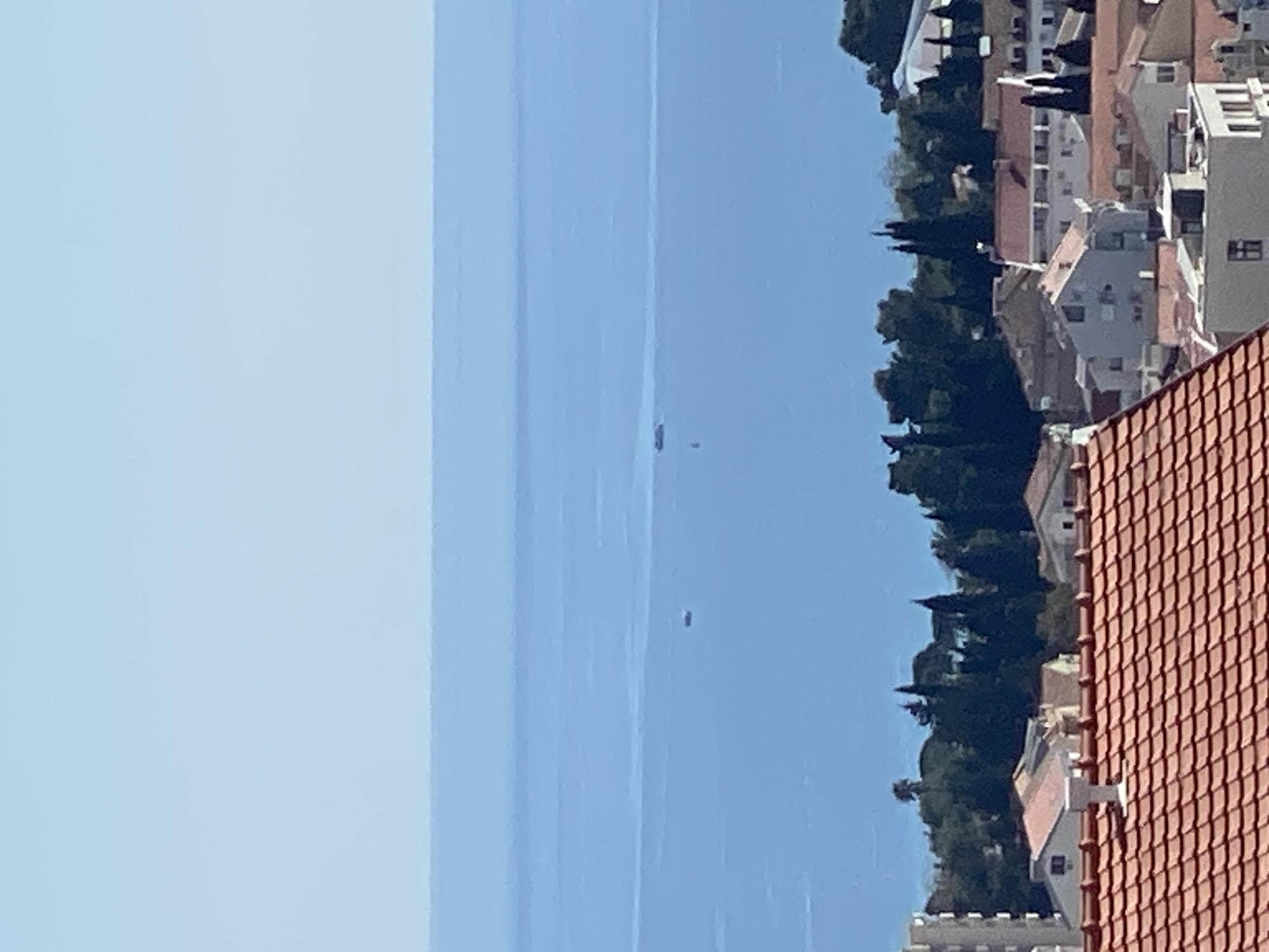 The image depicts a view from an elevated position in Budva, Montenegro, overlooking the Adriatic Sea. The sky is clear and light blue, fading slightly towards the horizon. The sea is a darker shade of blue, with what appear to be two small vessels visible in the distance. The right side of the image features a portion of the townscape, characterized by buildings with red-tiled roofs interspersed with greenery. A section of a roof with terracotta-colored tiles dominates the lower right corner. The buildings appear to be of varying heights and architectural styles. The overall scene conveys a bright, sunny day in a coastal town.