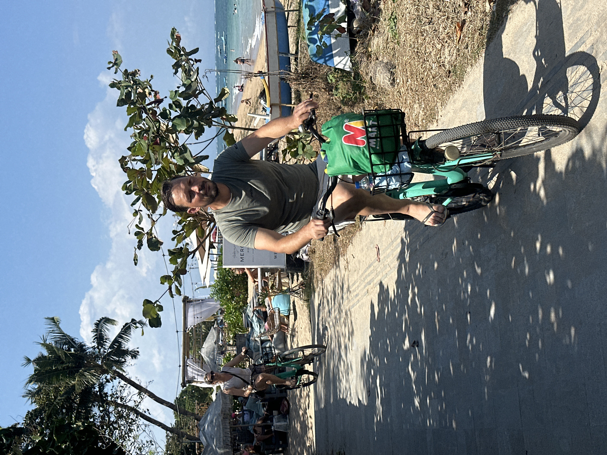The image depicts a sunny outdoor scene on a paved path near a beach. In the foreground, a man with short brown hair and a beard is riding a teal bicycle. He is wearing a green t-shirt and striped shorts. The bicycle has a green basket on the front containing a shopping bag with the red "M" logo of an Indonesian supermarket chain, "Matahari," along with a water bottle. In the background, a woman is also riding a teal bicycle. She is wearing sunglasses, a light-colored top, and shorts. Beyond the path, a beach is visible with lounge chairs, umbrellas, and people in the water. Palm trees and other foliage are scattered throughout the scene, and the ocean stretches to the horizon with several small boats visible. The overall impression is a casual, relaxed day at a beach resort. This scene is likely located in the Seminyak area of Denpasar, Indonesia, known for its beaches and vibrant atmosphere.