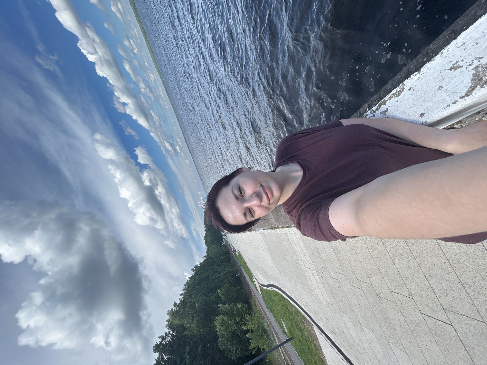 A woman with short dark hair and a maroon t-shirt takes a selfie outdoors on a paved walkway next to a large body of water. The walkway is made of large rectangular pavers and has a grassy area and trees to the left. The water is dark and choppy with whitecaps. The sky is blue with scattered white and gray clouds, and a distant tree line is visible across the water. The image appears to have been taken in Chaikovsky, Russia, based on the context provided.