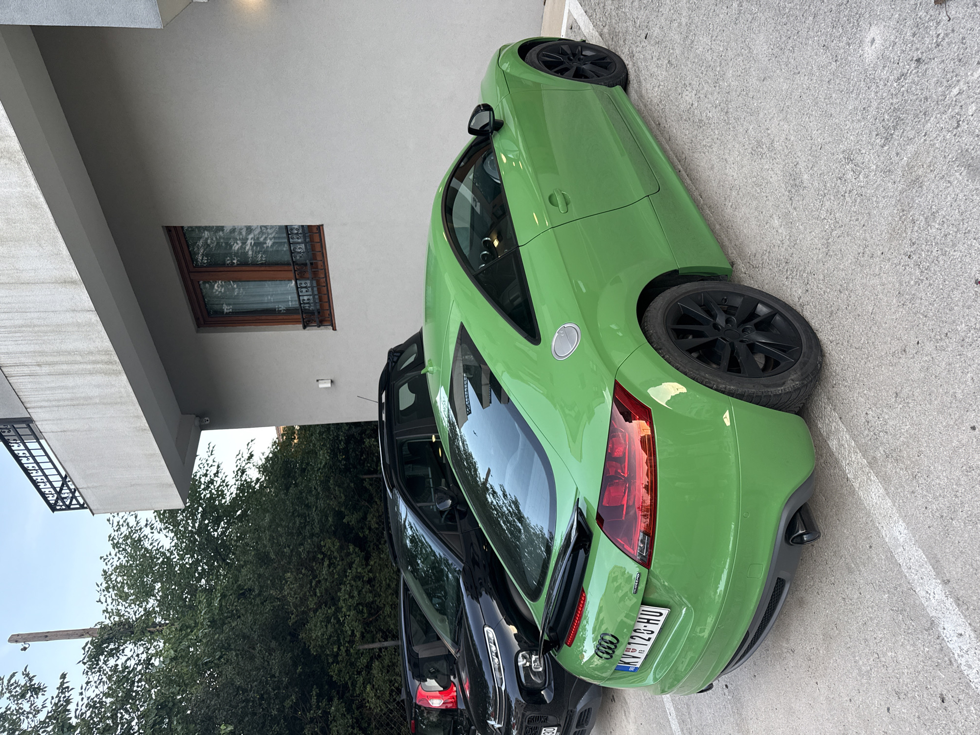 The image depicts a green Audi TT parked next to a black vehicle in what appears to be a parking lot in Budva, Montenegro. The license plate of the Audi TT is clearly visible, showing "KV 126-HU". Behind the cars are trees and part of a building with a window. The parking lot has concrete paving. The scene is captured during daylight.