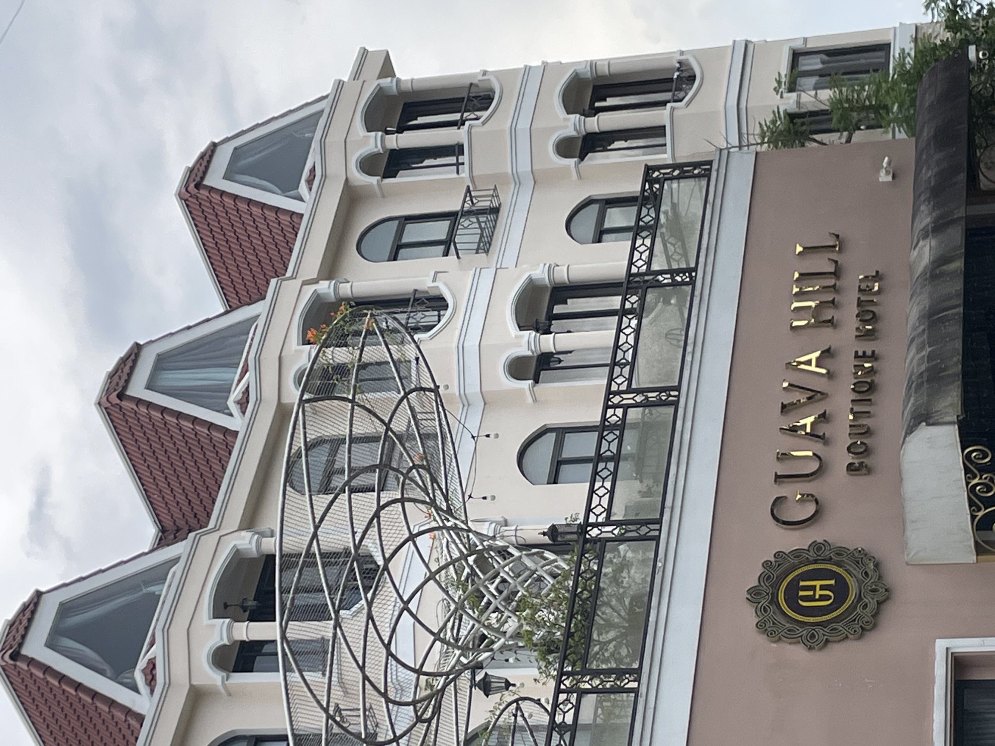 The image shows the exterior of the Guava Hill Boutique Hotel, located in Lâm Viên - Đà Lạt, Vietnam. The hotel is a multi-story building with a light beige facade and a dark red tiled roof. The architecture features arched windows, balconies with ornate railings, and decorative columns. A prominent sign on the lower part of the building reads "GUAVA HILL BOUTIQUE HOTEL" in gold lettering. A circular emblem with the letters "GH" is also visible next to the hotel name. The building is surrounded by greenery, with trees visible on the right side of the image. The sky is overcast.
