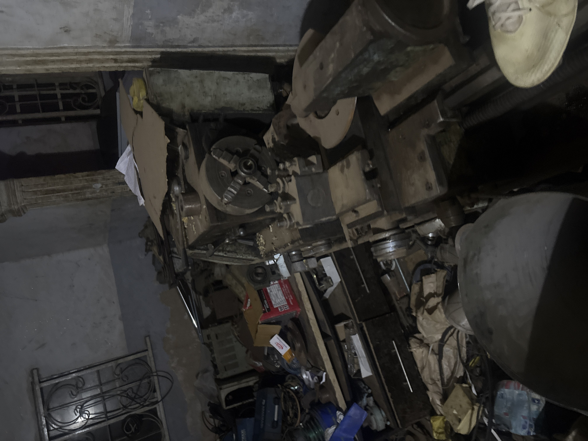 The image shows a cluttered indoor space in Bukuru, Nigeria. Dominating the scene is a large, industrial metal lathe. The surrounding area contains various tools, packaging, and miscellaneous objects scattered on the floor. A metal frame structure, possibly a bed frame or shelf, is visible in the background. The room is dimly lit and appears to be in a state of disrepair. A partially visible shoe is resting on the lathe.